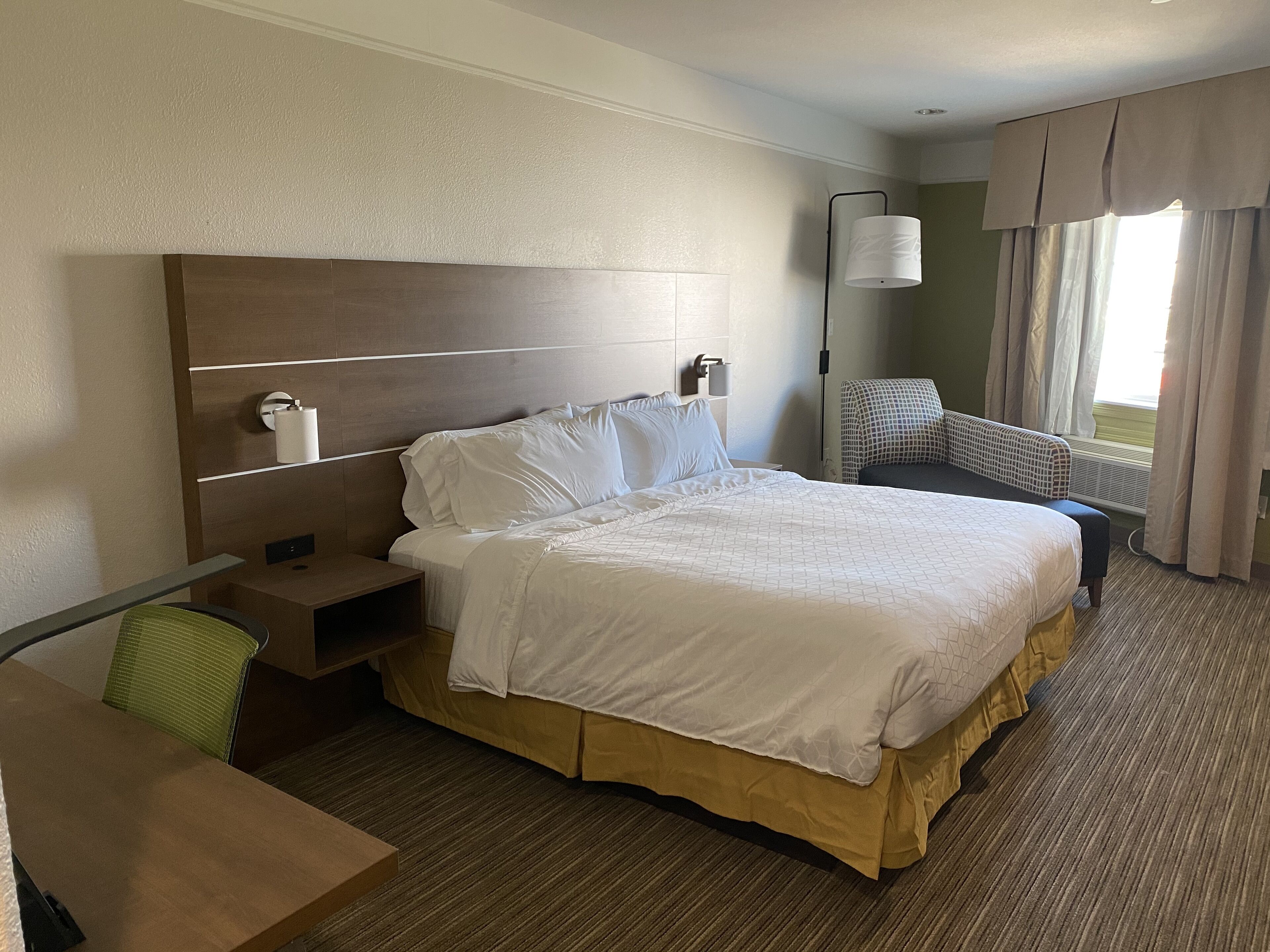 A hotel room with a large bed, a desk, a chair, a nightstand, and a lamp.