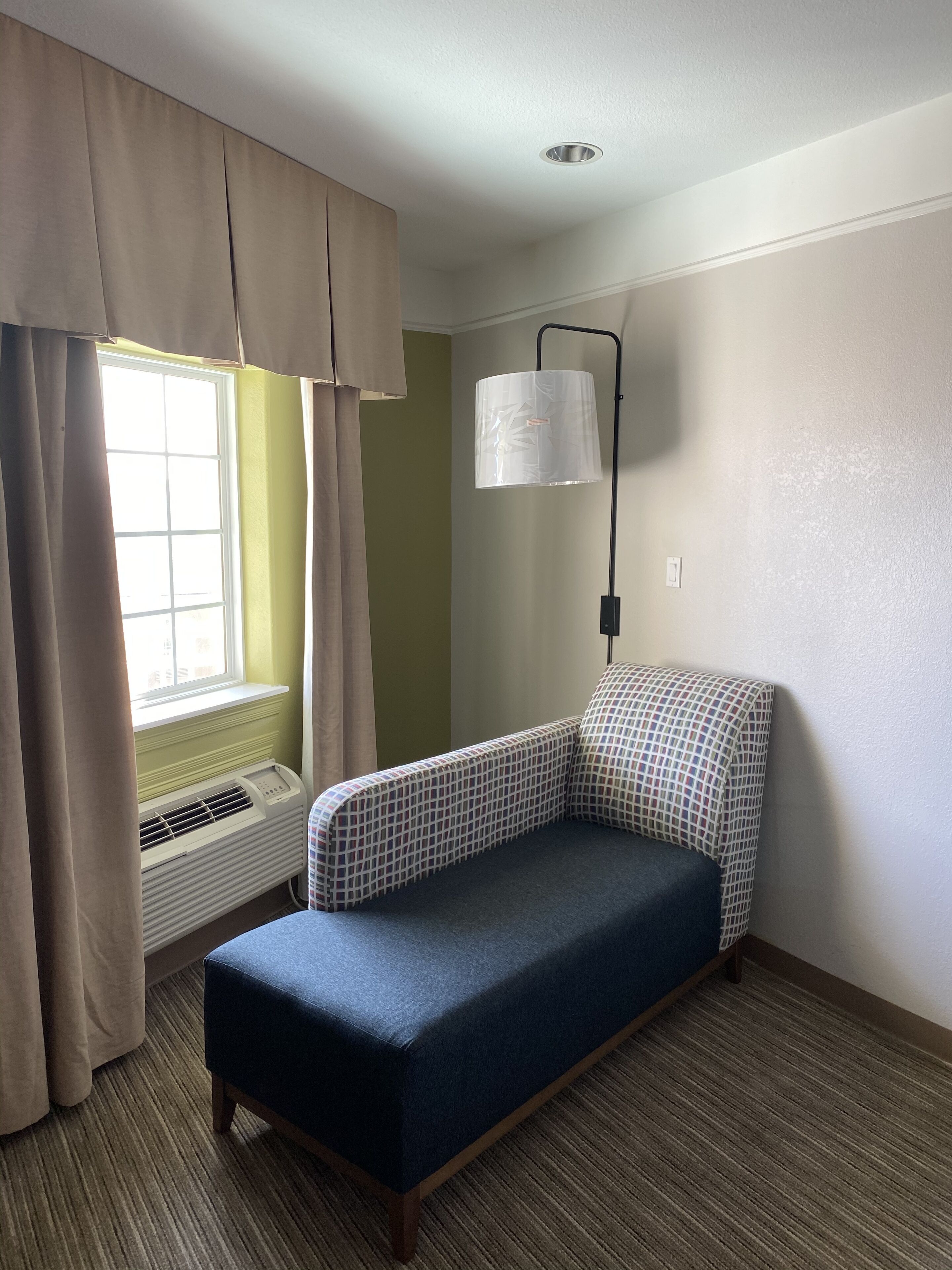 Een kleine kamer met een enkel bed, een raam, een airconditioner, een lamp en een hoofdbord met een patroon.