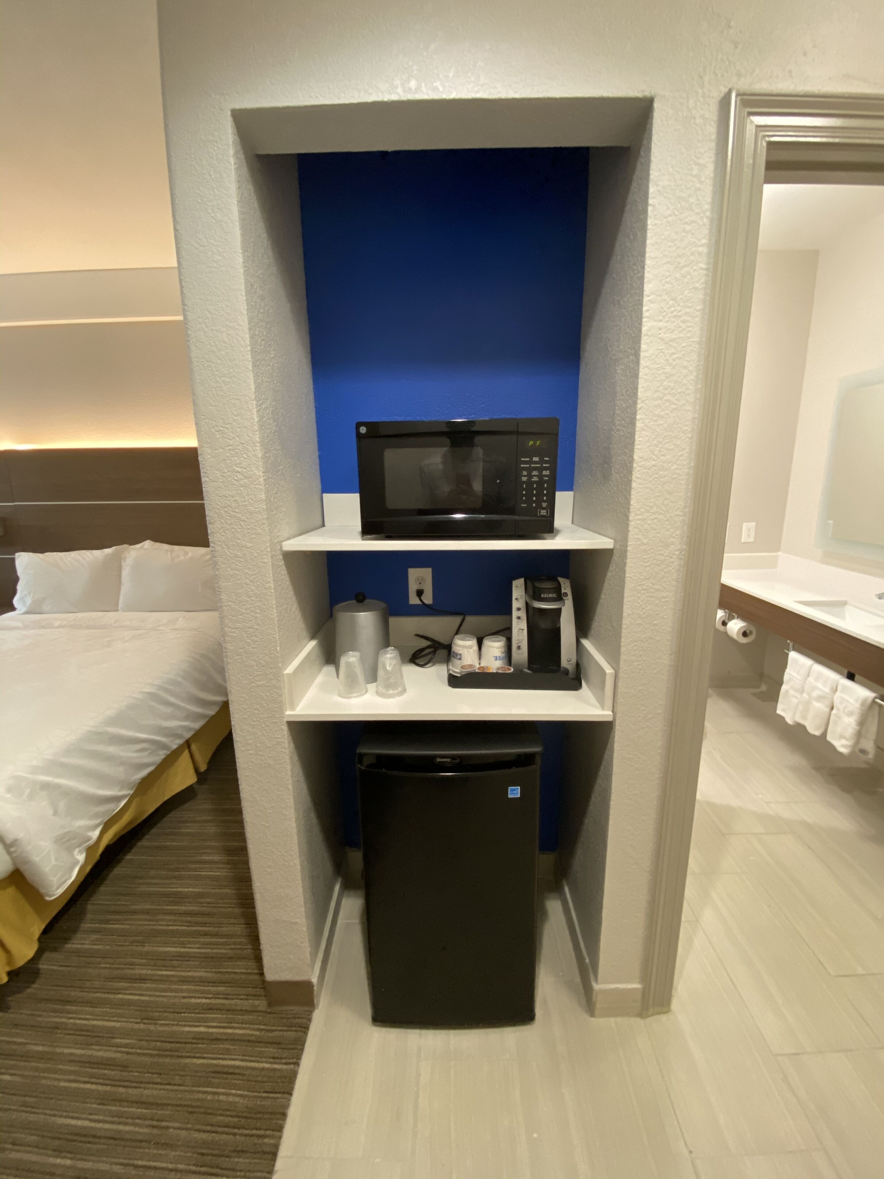 Habitación de hotel compacta con cama, microondas, cafetera y refrigerador pequeño.