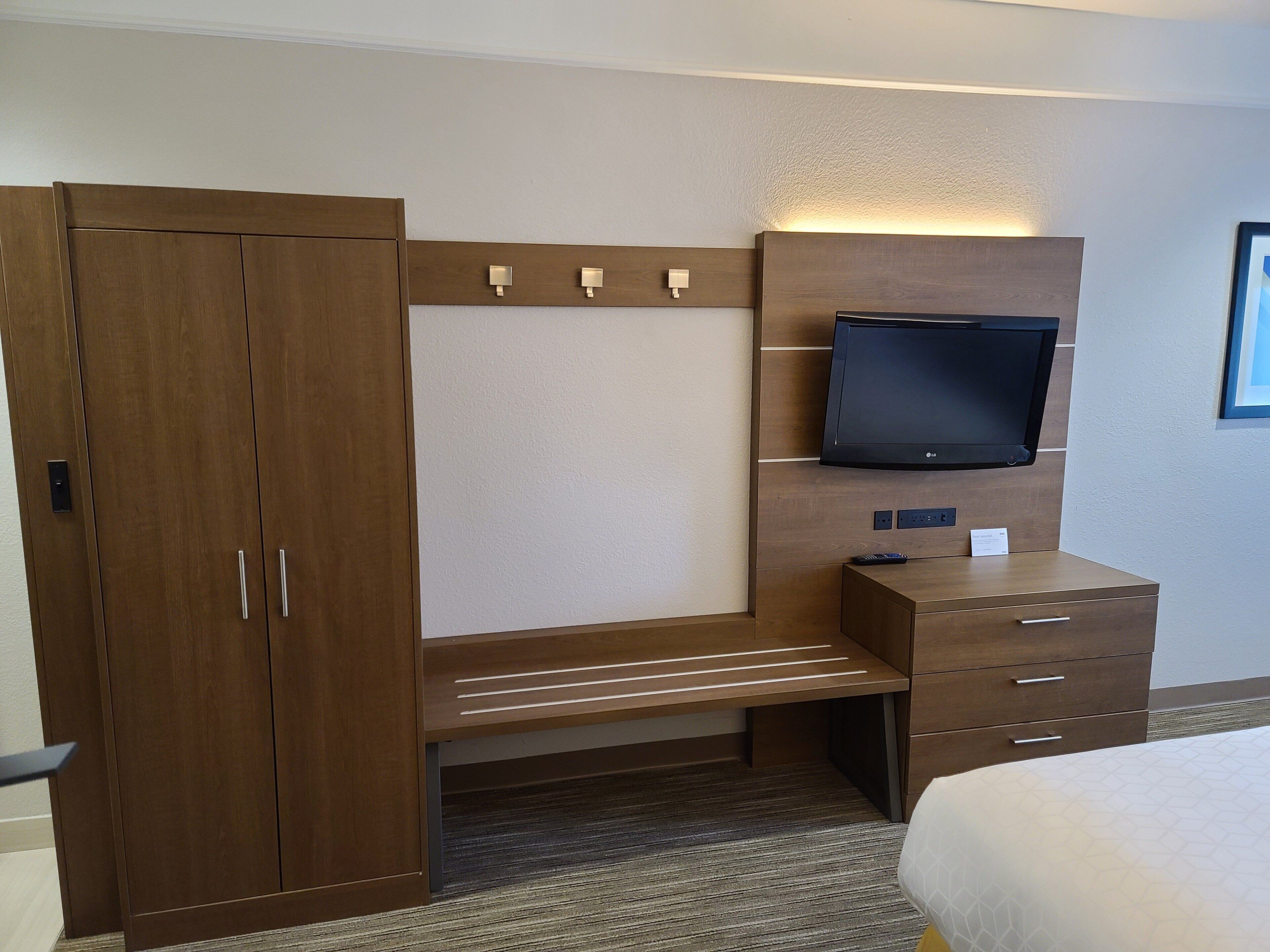 A hotel room with a wooden wardrobe, a desk with drawers, a mounted flat-screen TV, and a bed with a patterned bedspread.