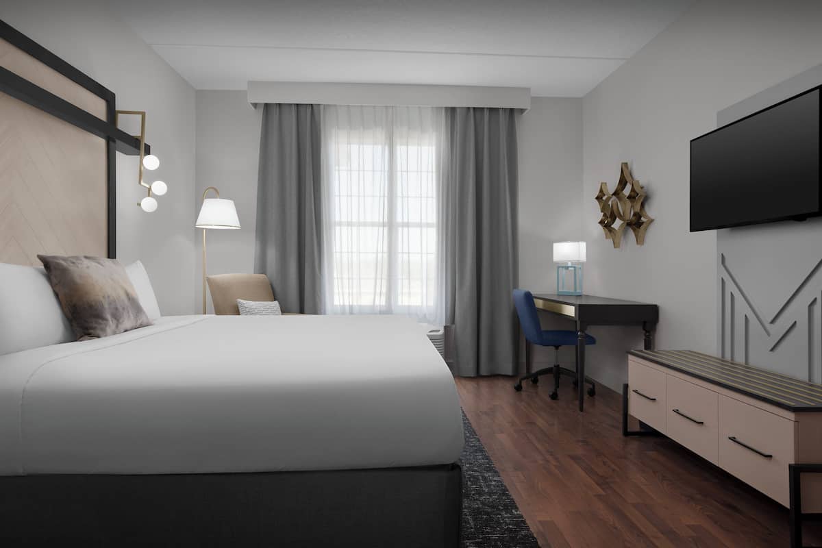 A modern hotel room with a large bed, a desk, a chair, a TV, and a desk lamp.