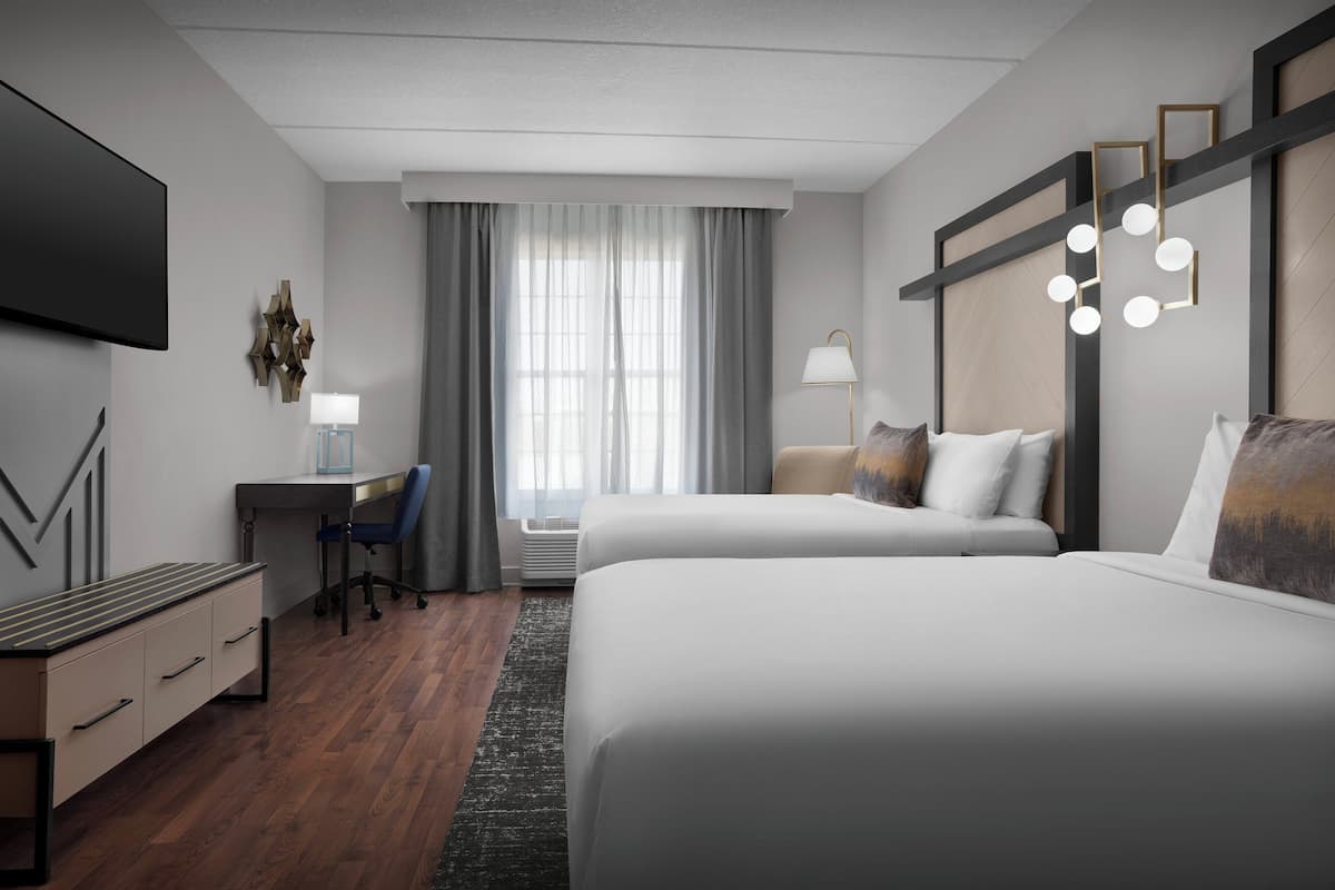 A hotel room with two beds, a desk, a chair, a TV, and a large window with curtains.