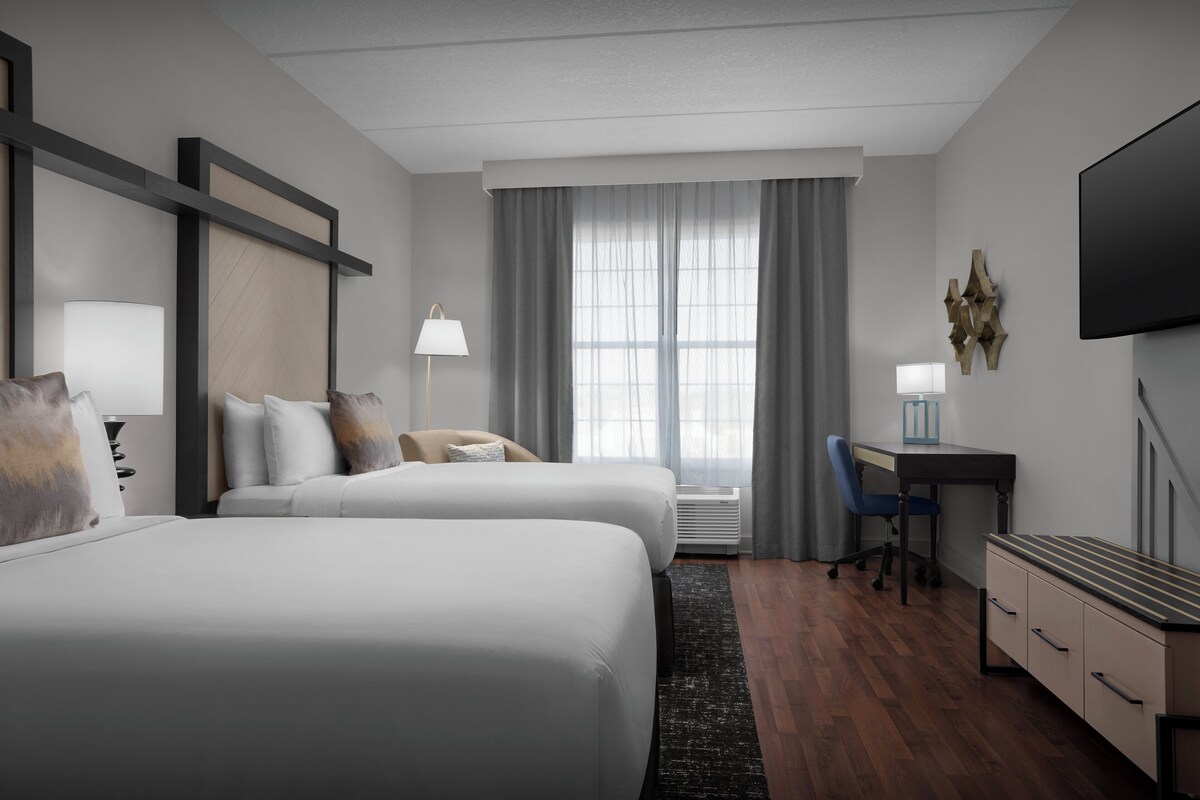 Une chambre d’hôtel avec deux lits, un bureau, une chaise, une télévision et une fenêtre avec des rideaux.