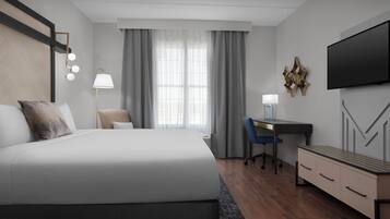A modern hotel room with a large bed, a desk, a chair, a TV, and a desk lamp.