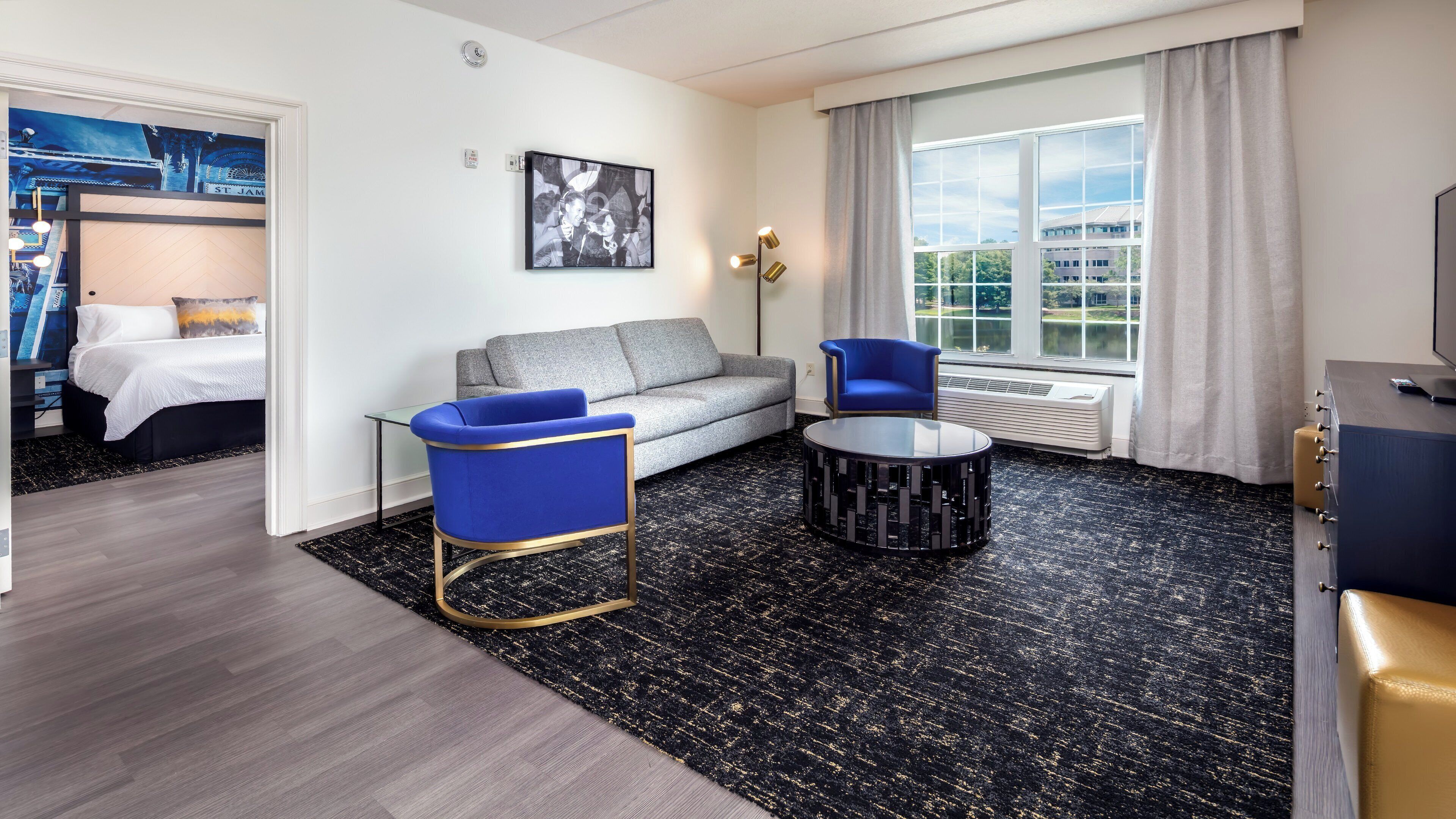 A modern hotel room with a grey sofa, a blue armchair, a black and gold coffee table, and a large window with a view of greenery.