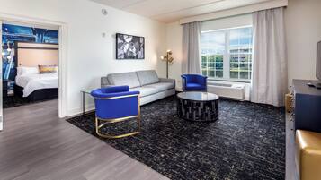 A modern hotel room with a grey sofa, a blue armchair, a black and gold coffee table, and a large window with a view of greenery.