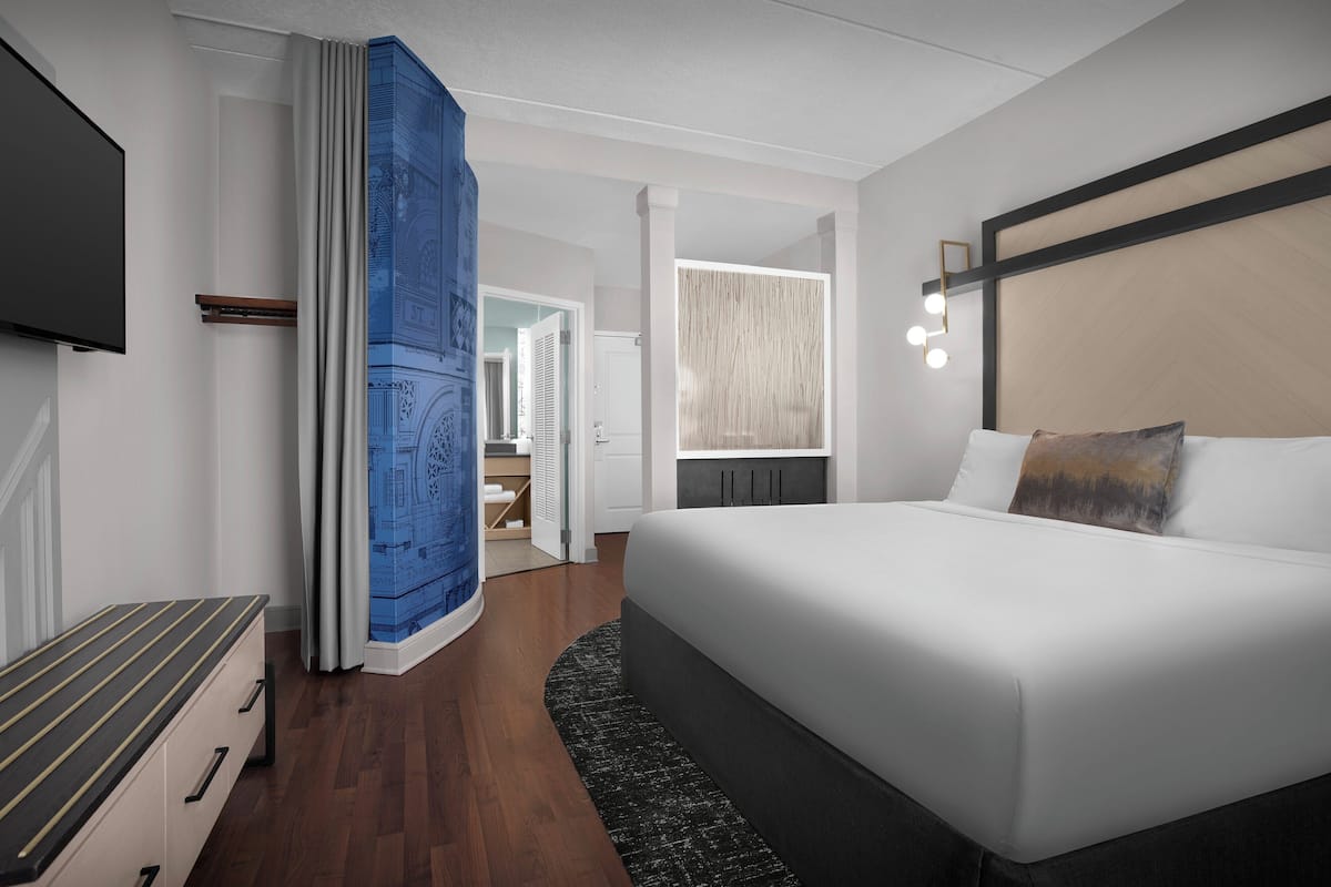 A modern hotel room with a large bed, a flat-screen TV, a desk, and a view of the hallway.