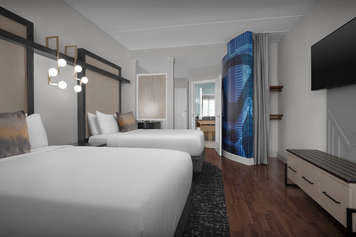 A modern hotel room with two beds, a flat-screen TV, a desk, and a large window with a view of a building.