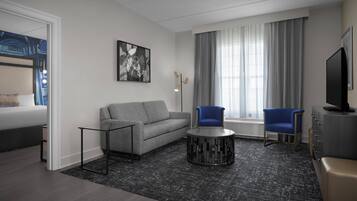 Ein modernes Hotelzimmer mit grauem Sofa, zwei blauen Sesseln, einem runden schwarzen Couchtisch, einem Flachbildfernseher und einem großen Fenster mit Vorhängen.