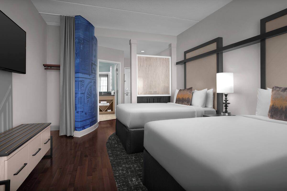 A hotel room with two beds, a TV, a desk, and a bathroom visible through an open door.