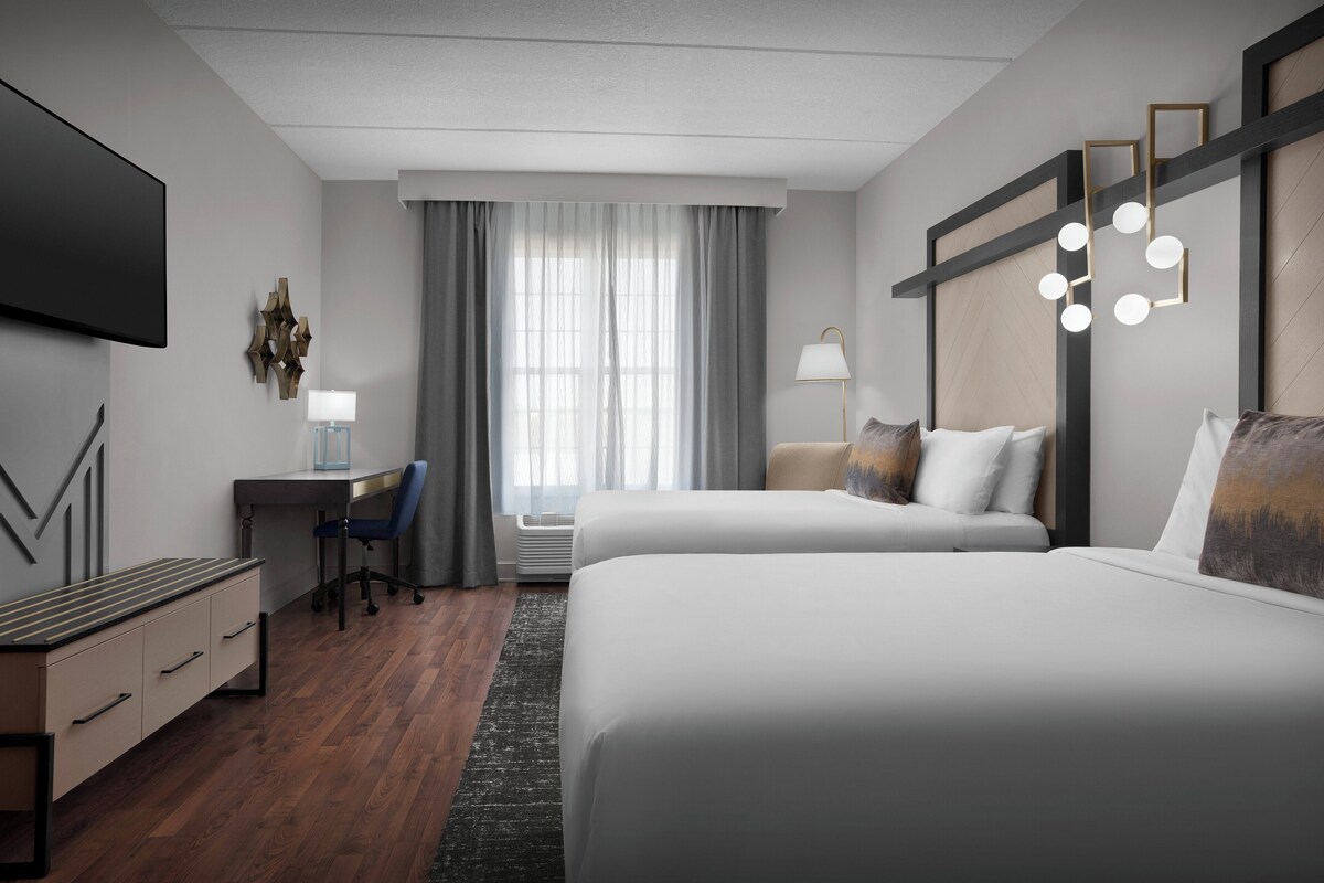 Une chambre d’hôtel avec deux lits, un bureau, une chaise, une télévision et une grande fenêtre avec des rideaux.