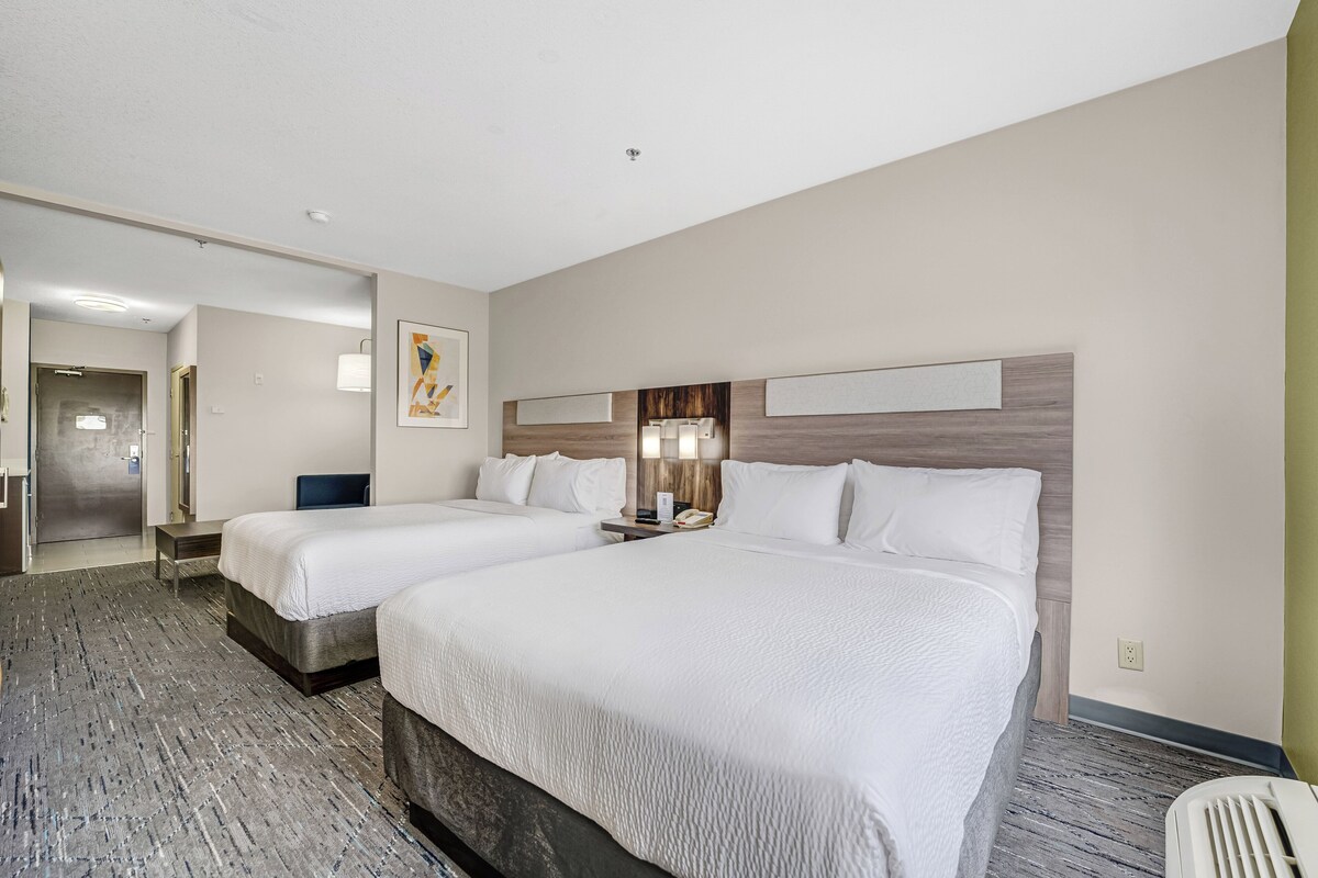 A hotel room with two beds, a desk, a chair, a lamp, and a wall-mounted TV.