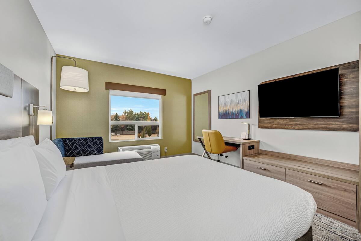 A modern hotel room with a large bed, a desk, a chair, a flat-screen TV, and a window with a view of trees.