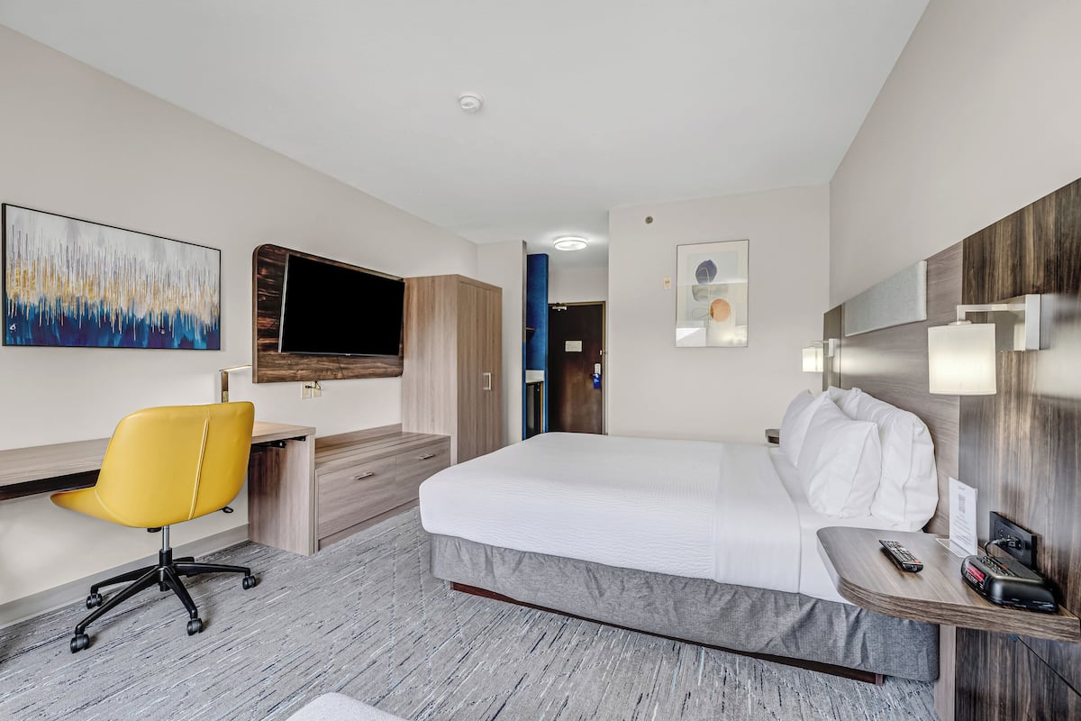 A modern hotel room with a bed, a desk with a chair, a flat-screen TV, and artwork on the walls.