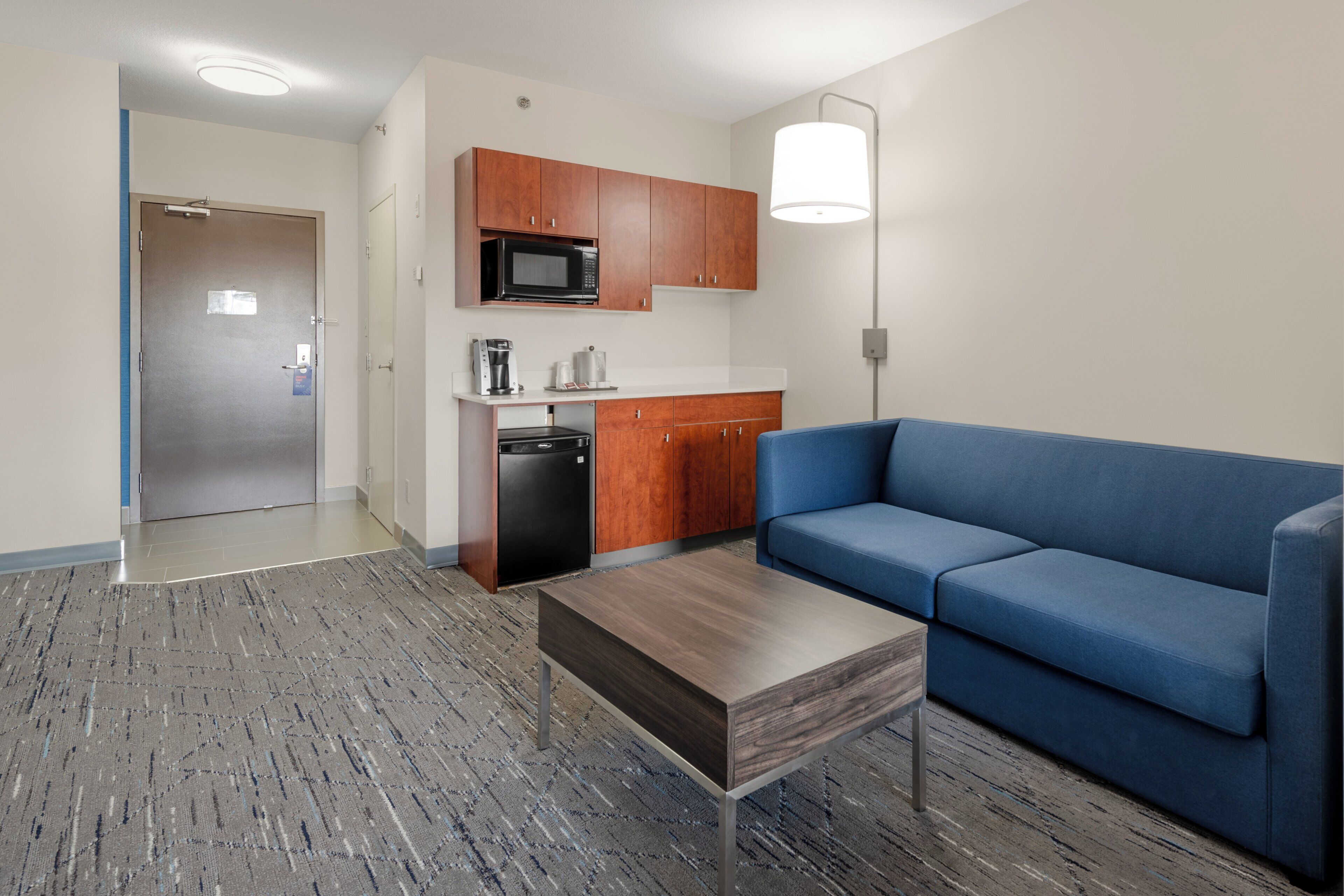 A modern living room with a blue sofa, a wooden coffee table, and a kitchenette with a microwave.