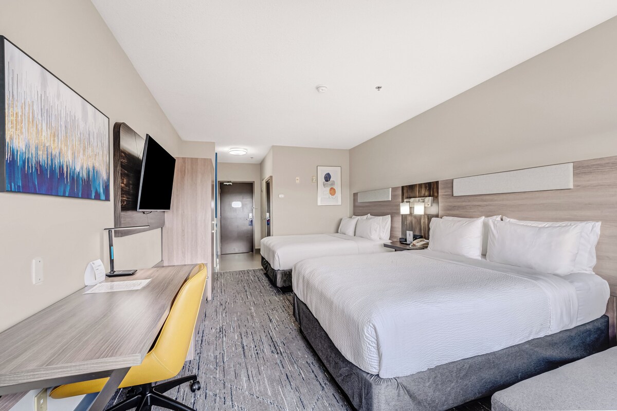 A hotel room with a desk, two beds, a TV, and a painting on the wall.