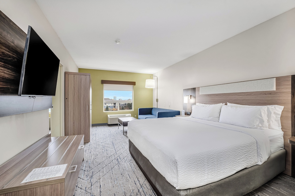 Une chambre d’hôtel avec un grand lit, un téléviseur à écran plat fixé au mur, un bureau, une chaise et une fenêtre donnant sur des immeubles.
