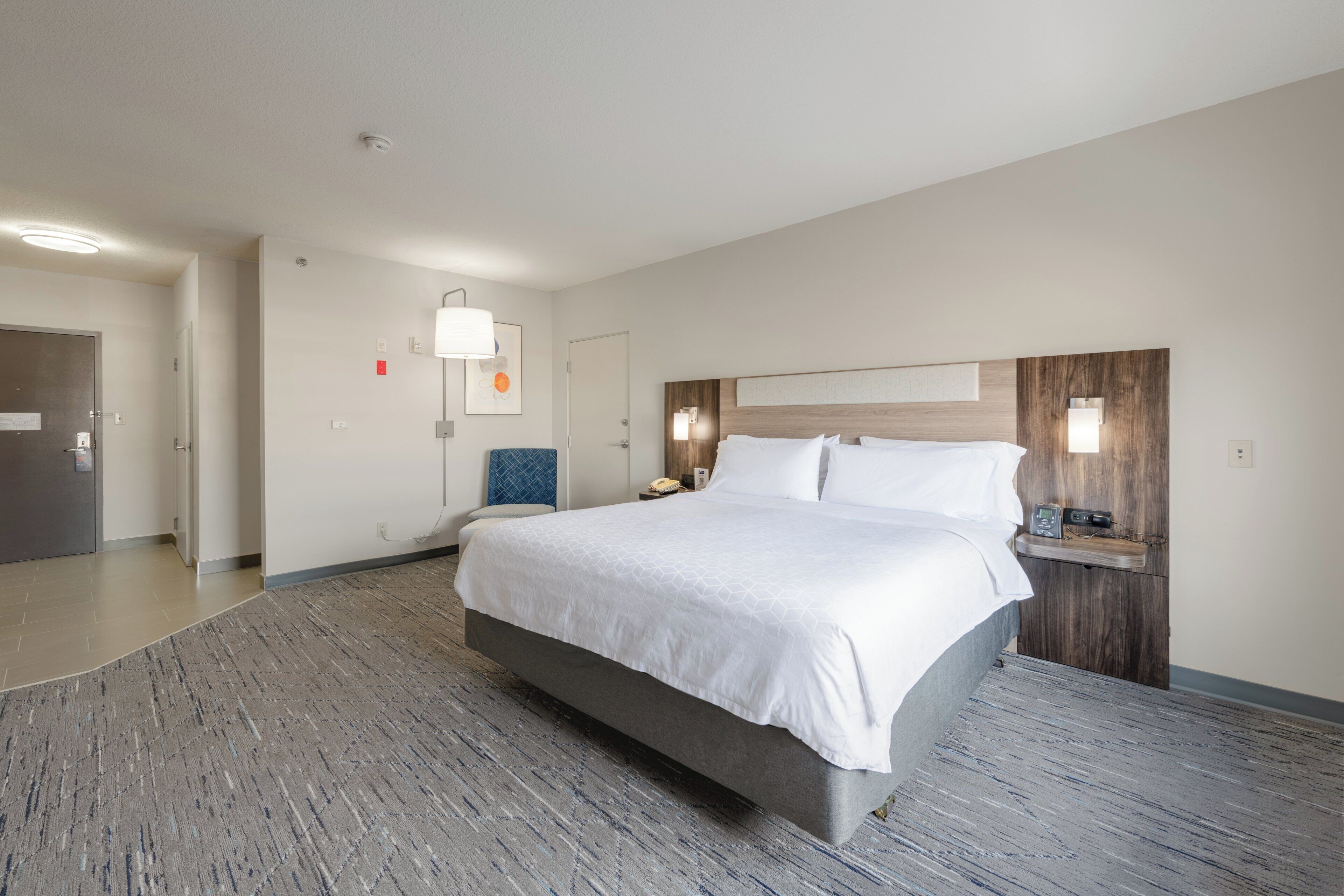 A hotel room with a large bed, a bedside table with a phone, a wall-mounted light, and a door leading to another room.