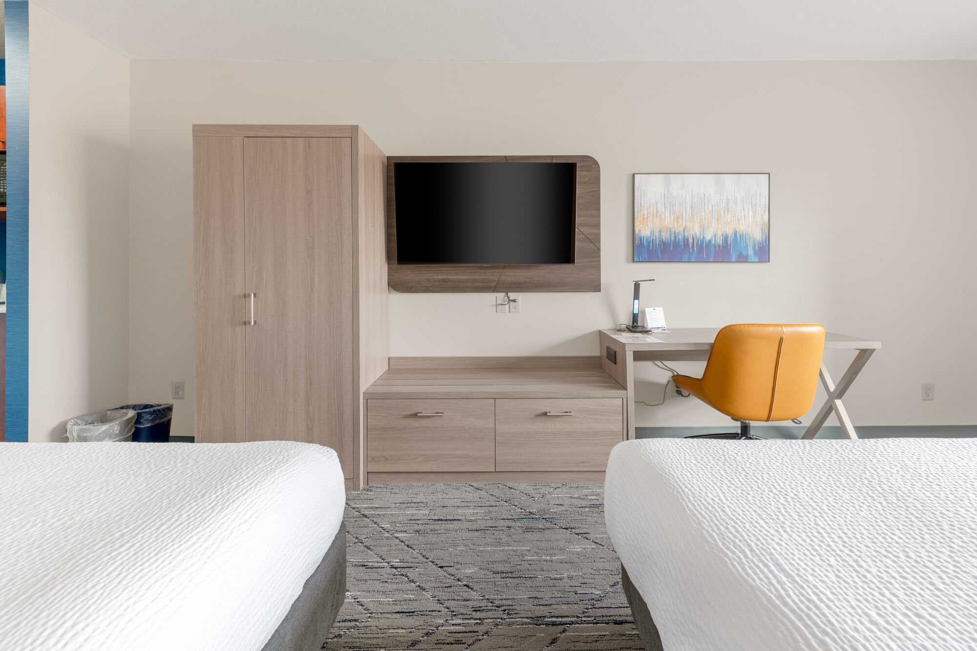 A hotel room with two beds, a desk with a chair, a TV mounted on the wall, and a painting above the desk.
