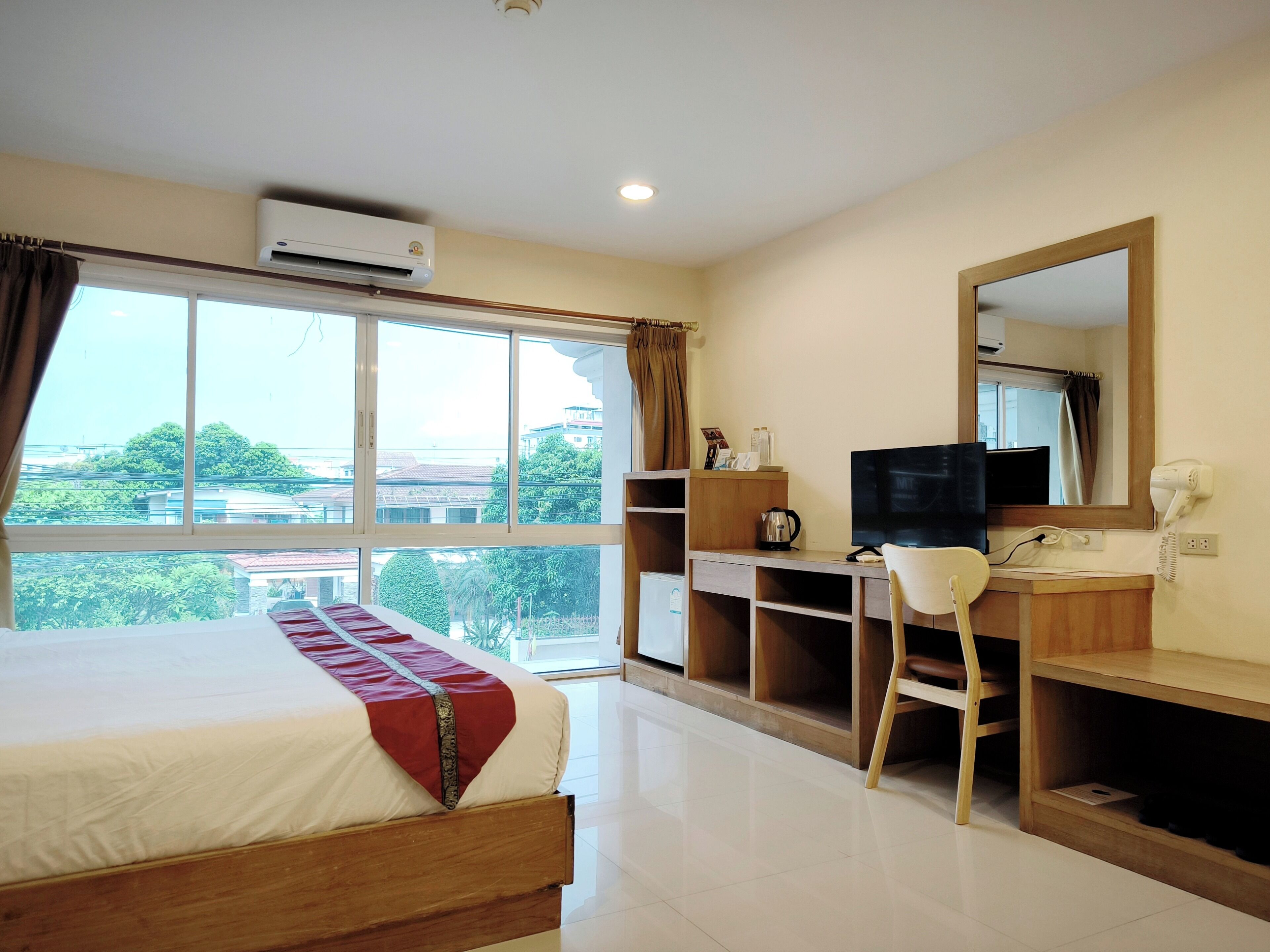 A hotel room with a bed, a desk with a chair, a television, an air conditioner, and a large window offering a view of buildings and greenery.