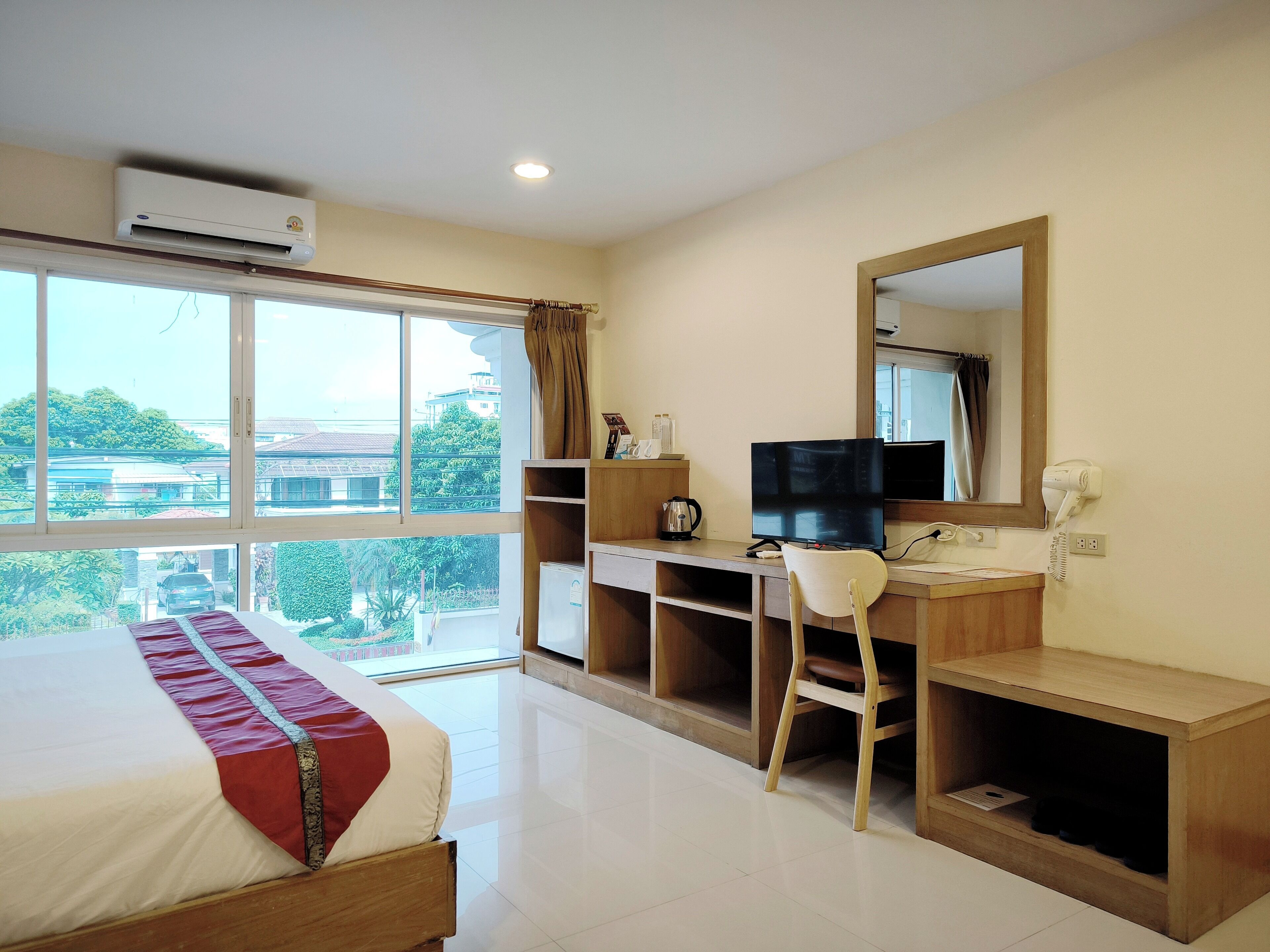 A hotel room with a bed, a desk with a computer, a TV, and a large window offering a view of buildings and trees.