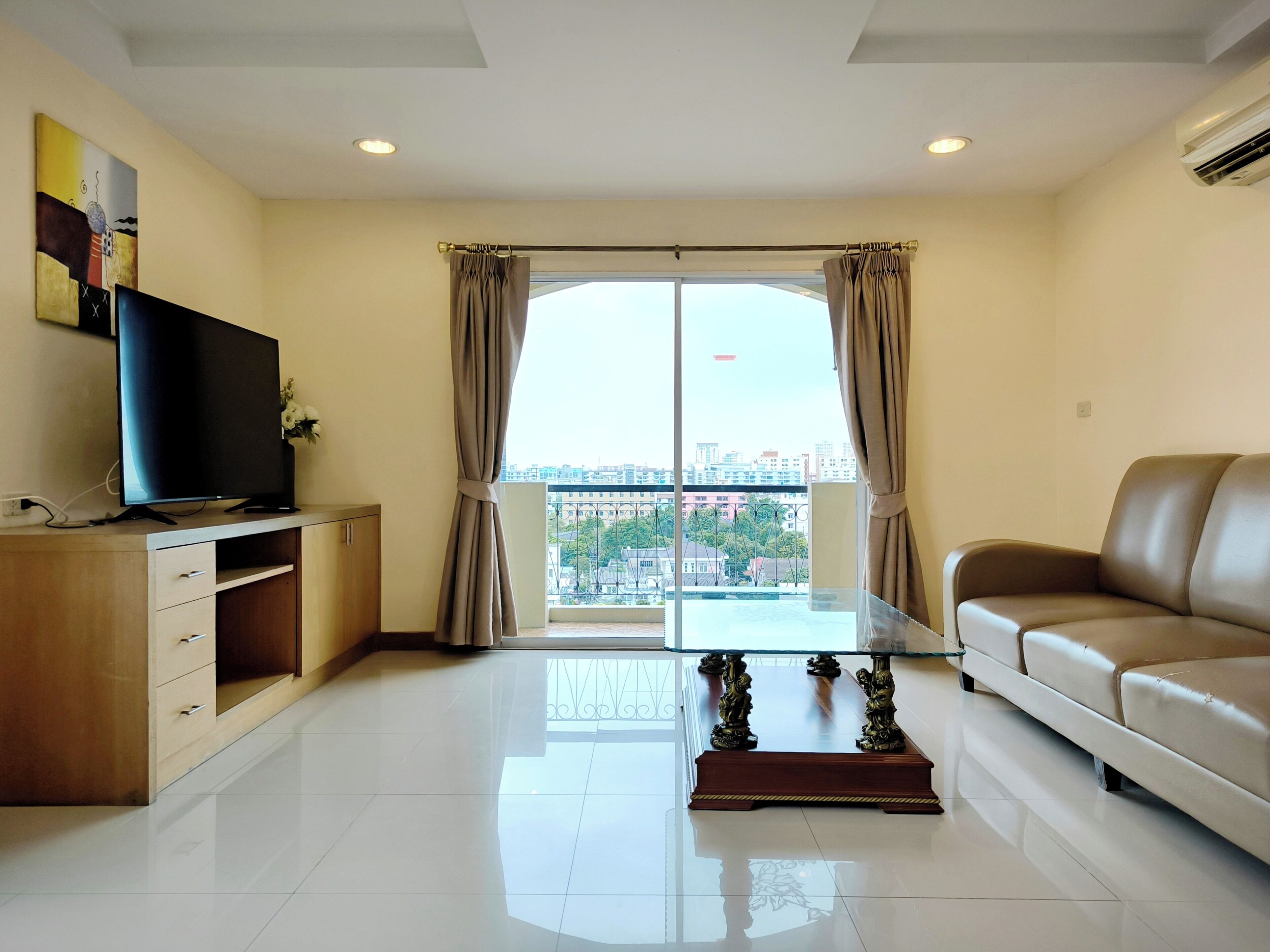 A modern living room with a flat-screen TV on a wooden cabinet, a glass-top coffee table with ornate legs, a beige sofa, and a large window with curtains.