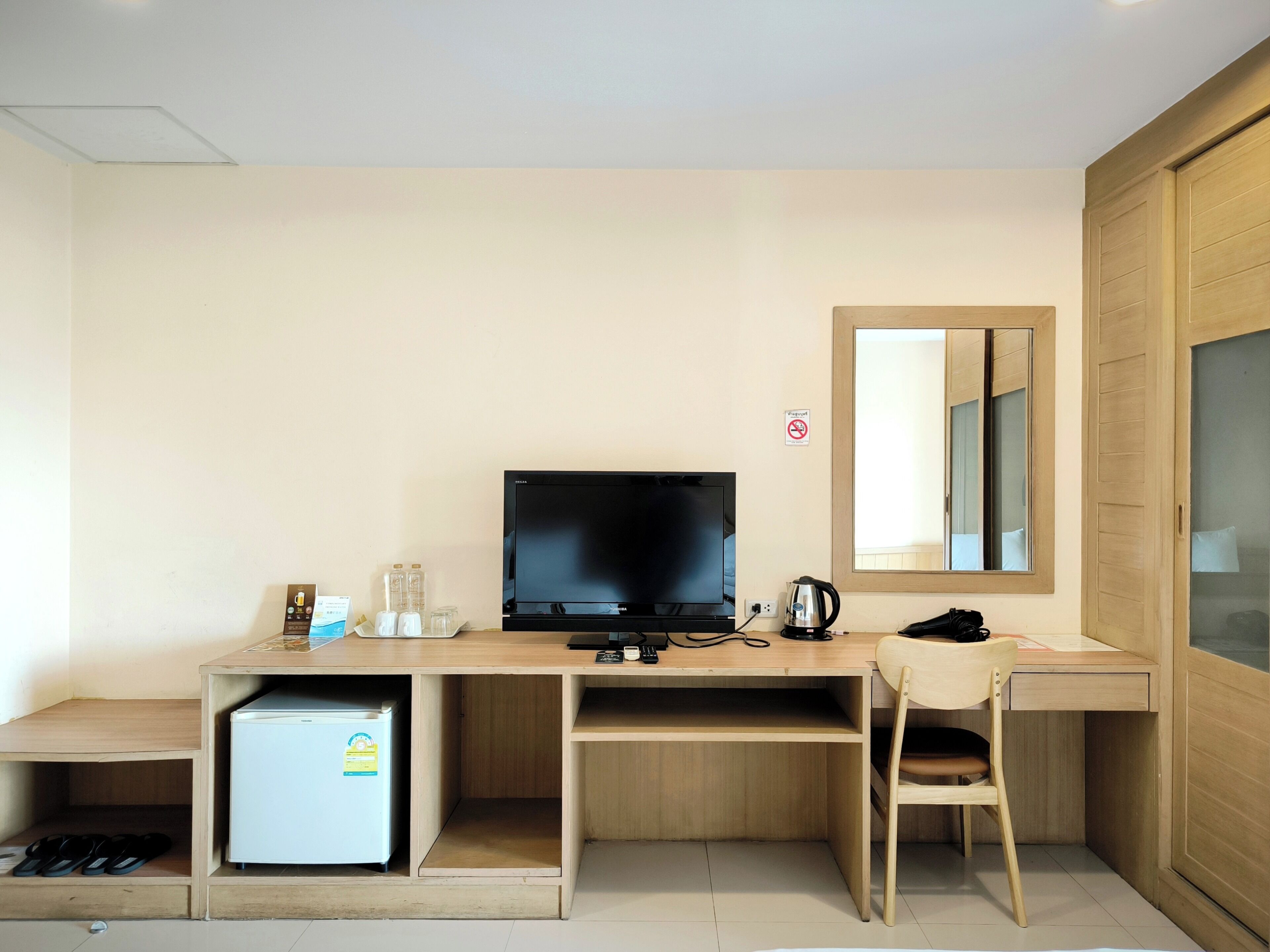 A compact hotel room with a wooden desk, a television, a small refrigerator, a chair, and a mirror.