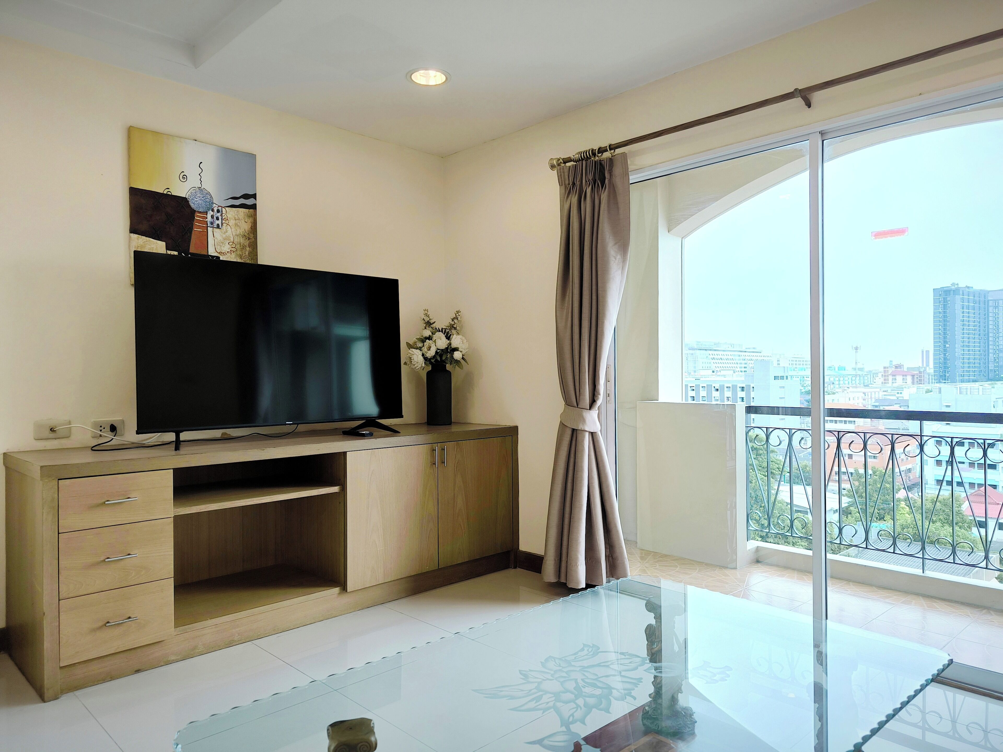 A modern living room with a flat-screen TV, wooden cabinet, glass coffee table, and a view of the city through large windows.