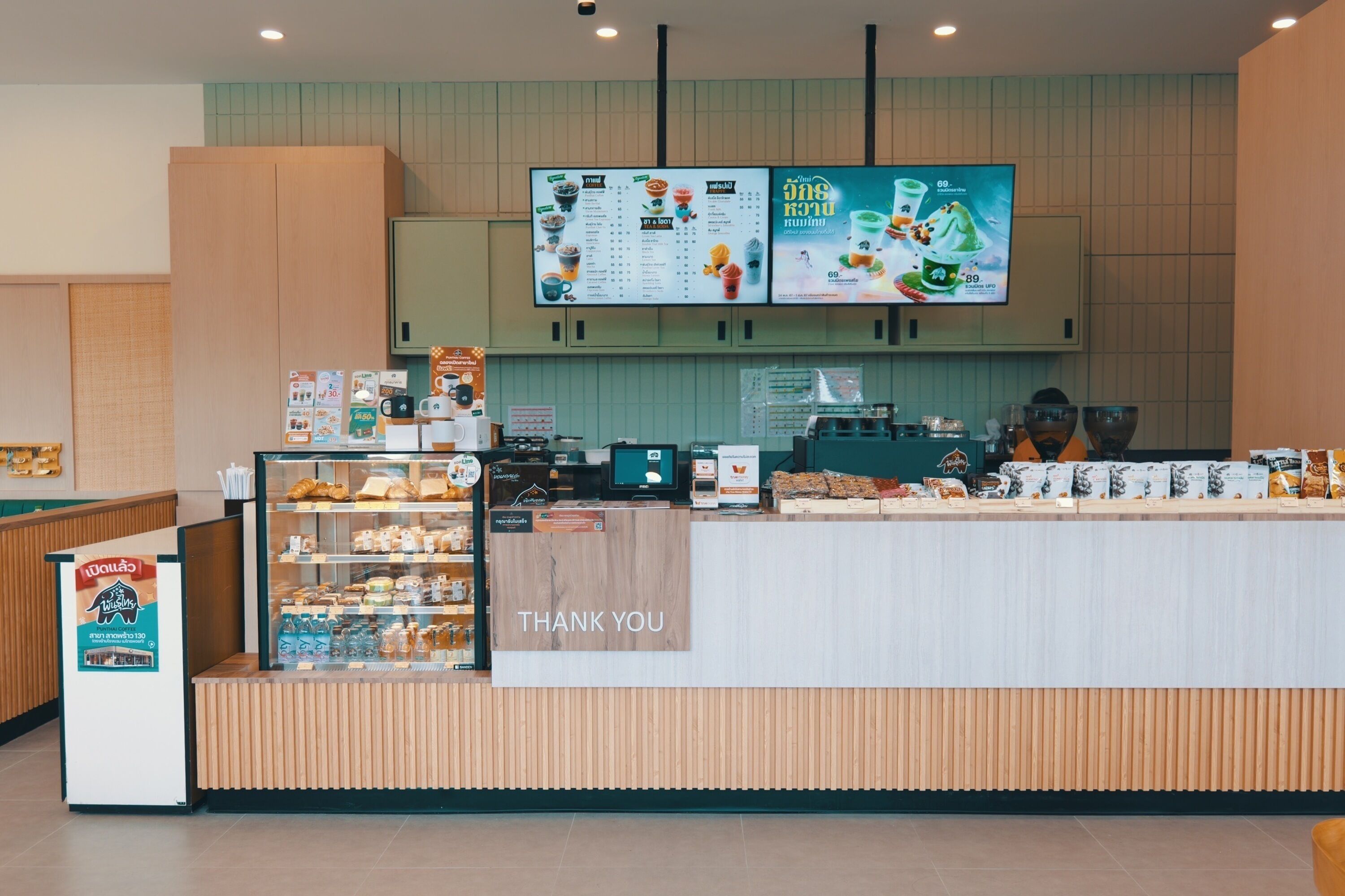 L’intérieur d’un café moderne présente un comptoir d’accueil en bois, des panneaux affichant le menu et une variété de produits alimentaires exposés.