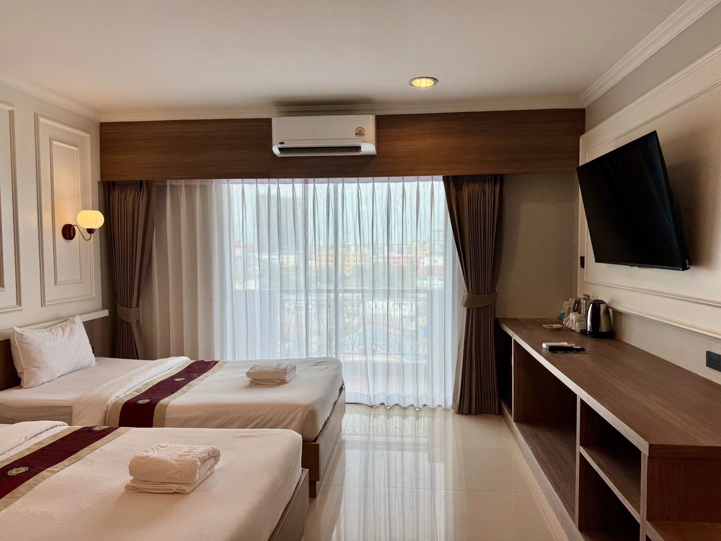A hotel room with two beds, a TV, a desk, and an air conditioning unit.