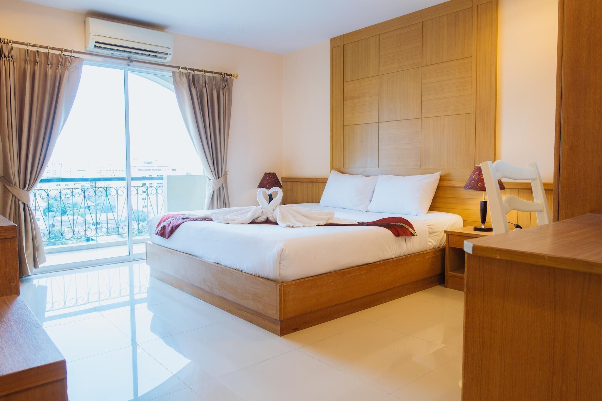 A hotel room with a wooden bed, a nightstand, a chair, and a view of the city through large windows.