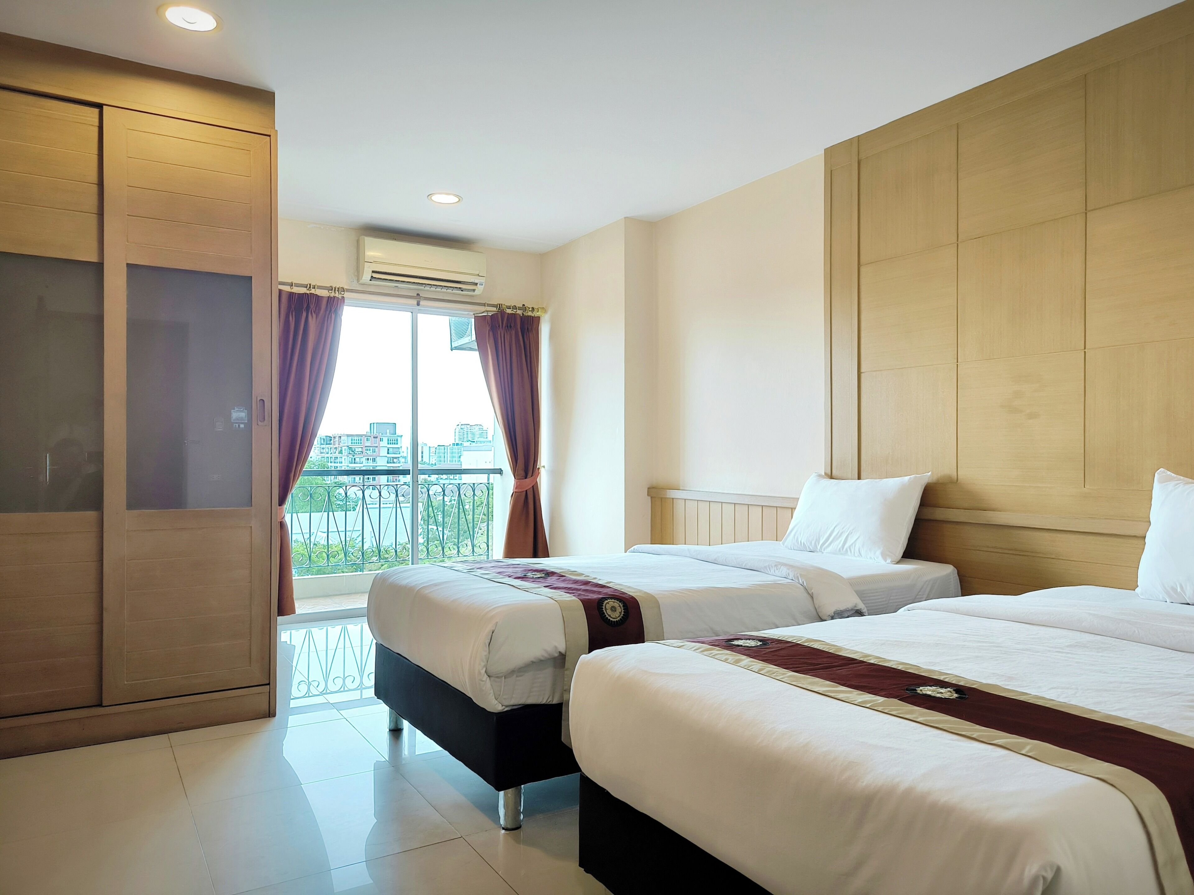 A hotel room with two beds, a large window with curtains, an air conditioning unit, and a balcony with a view of buildings.