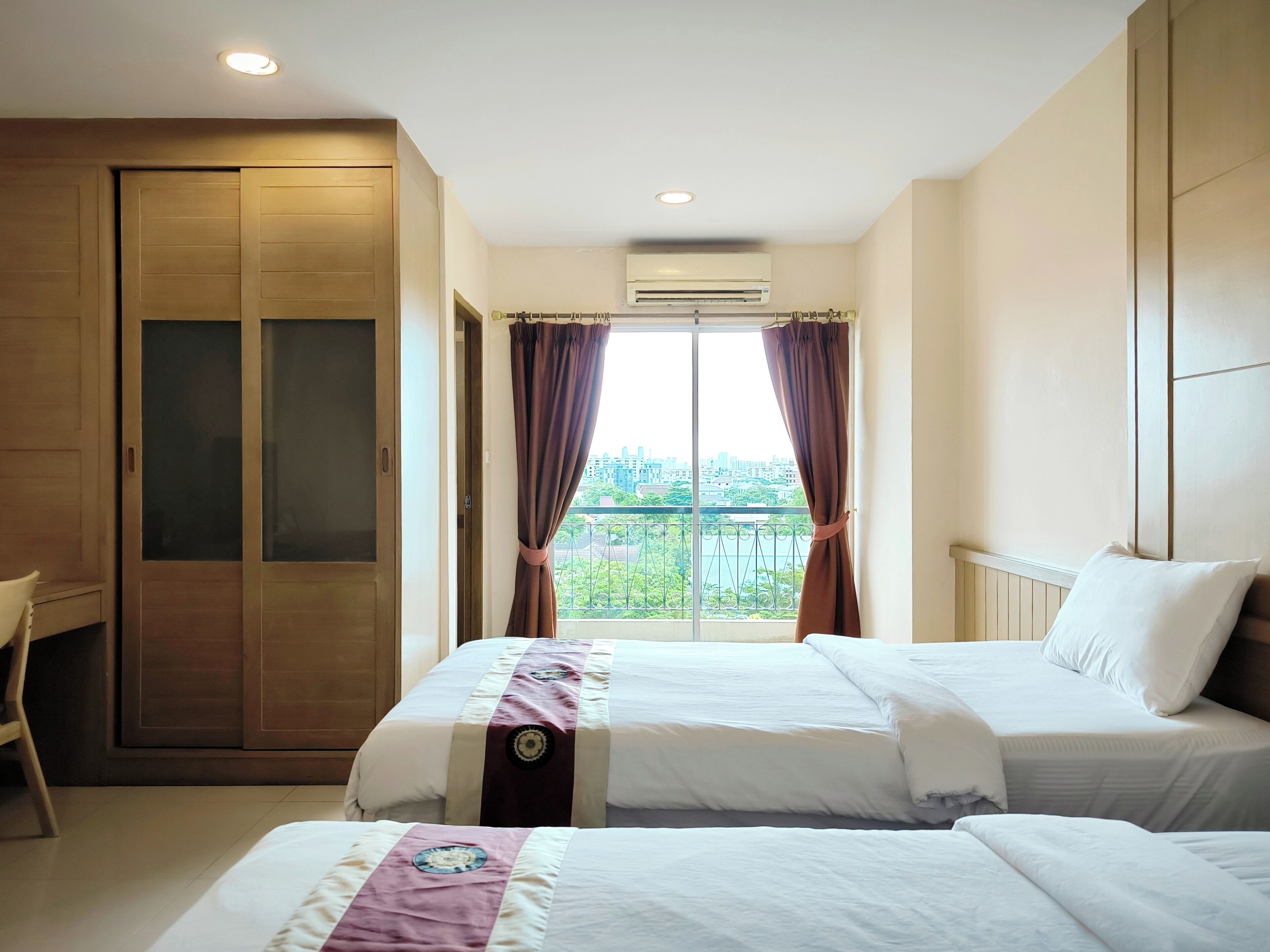 Habitación de hotel con dos camas, un escritorio y vistas a un paisaje urbano a través de un ventanal amplio.