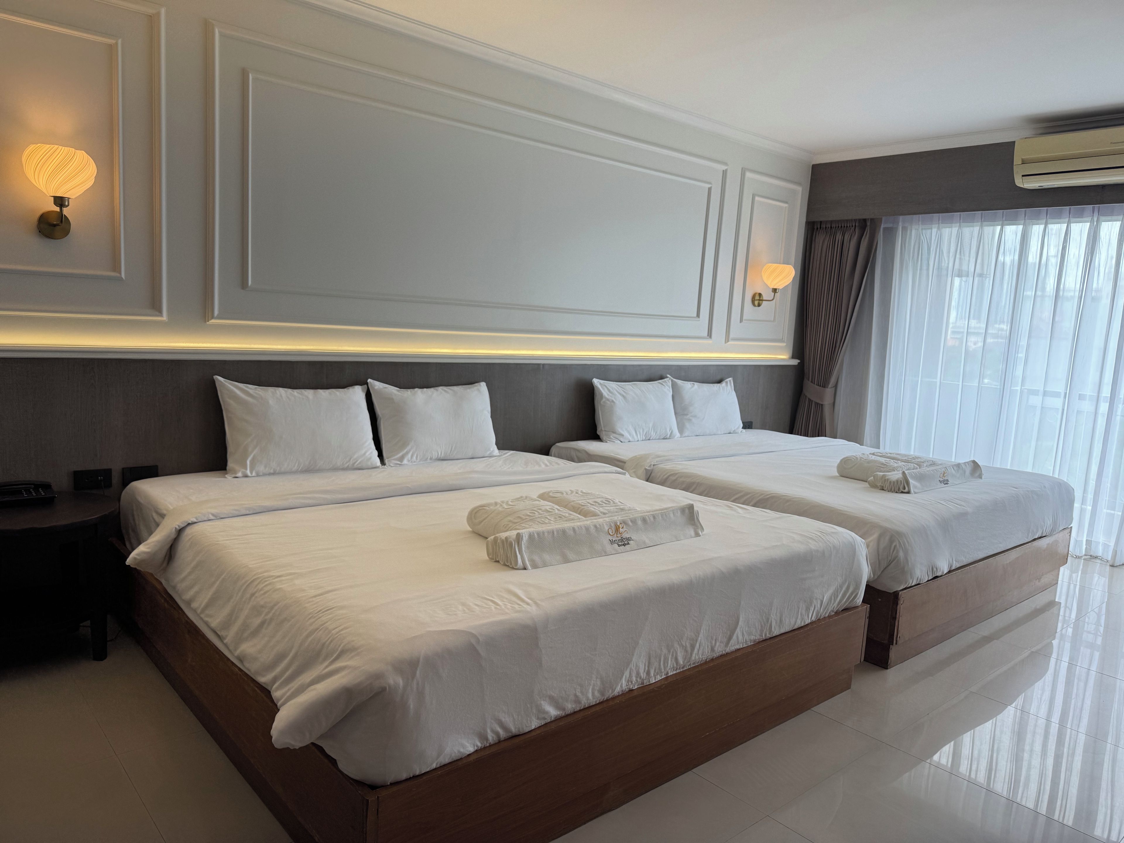 Habitació d'hotel amb dos llits, una taula de nit, una làmpada i una finestra gran amb cortines.