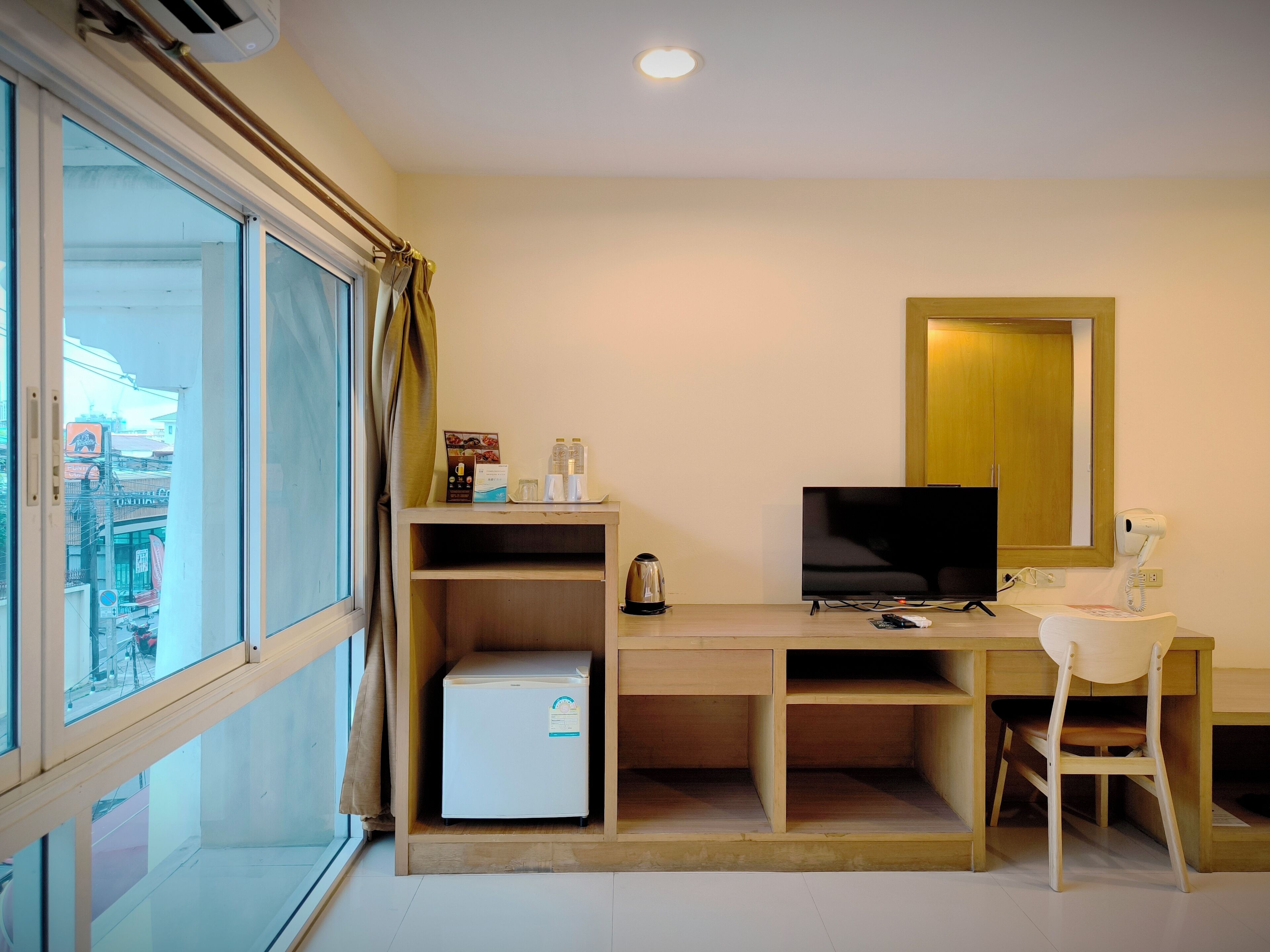 A hotel room with a desk, a television, a chair, a small refrigerator, and a window with curtains.