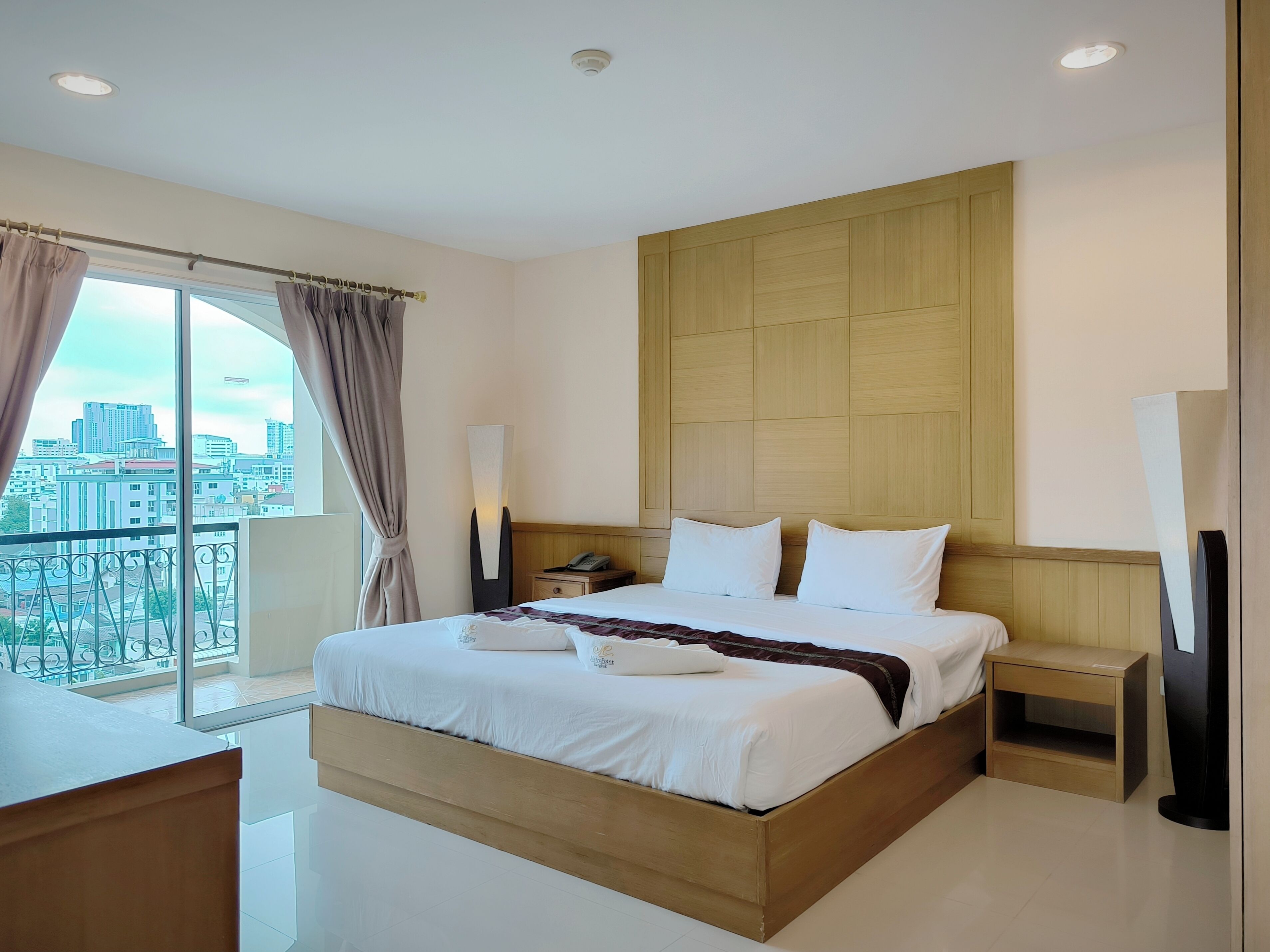 A modern hotel room with a large bed, a bedside table, a nightstand, a lamp, and a view of the city through a large window.
