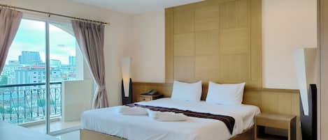 A modern hotel room with a large bed, a bedside table, a nightstand, a lamp, and a view of the city through a large window.