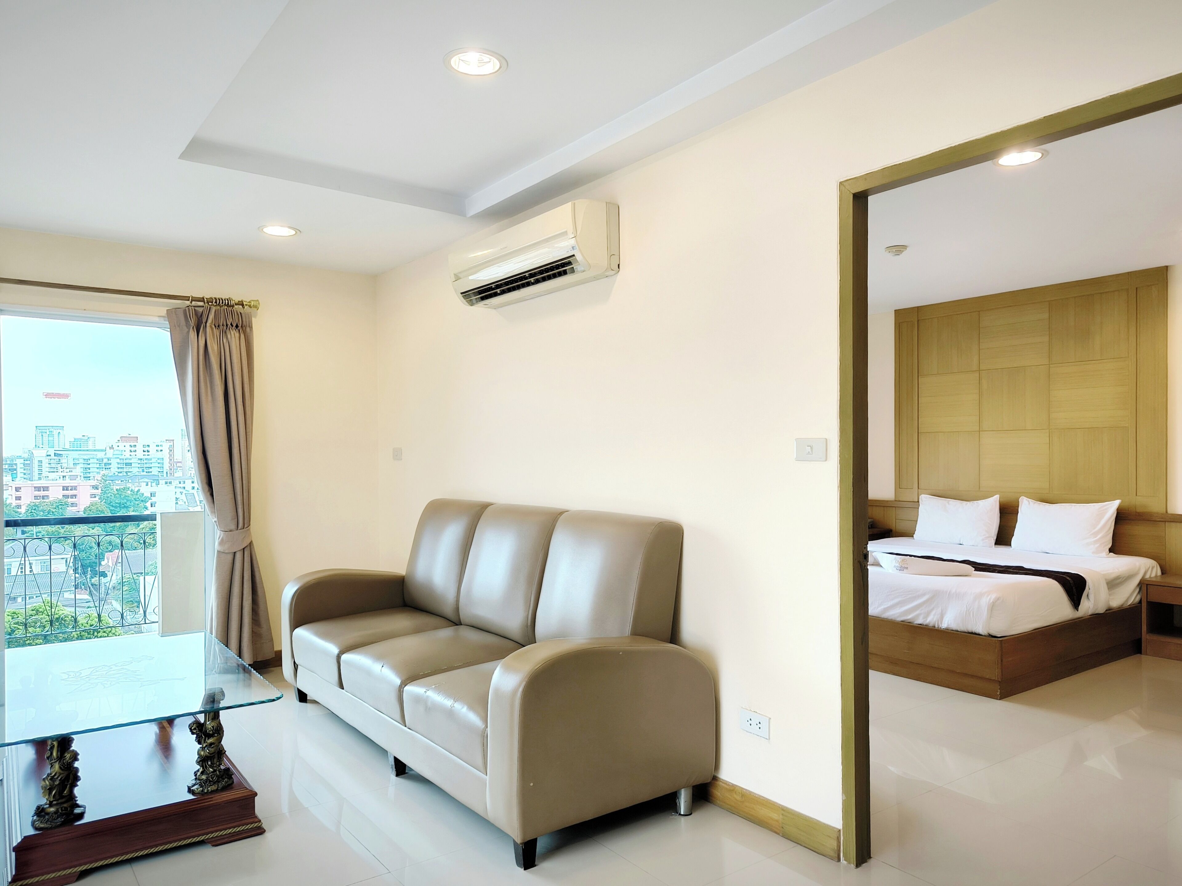 A modern hotel room with a leather sofa, a glass-top table, a bed with wooden headboard, and a city view through the window.