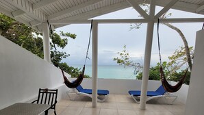 Uma varanda com vista para o mar, duas cadeiras de descanso azuis, uma mesa e uma rede.