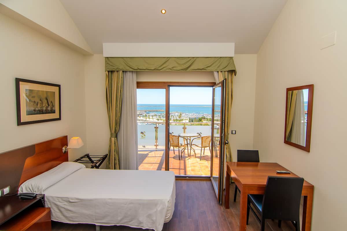 A hotel room with a bed, a desk, and a balcony with a view of the sea.