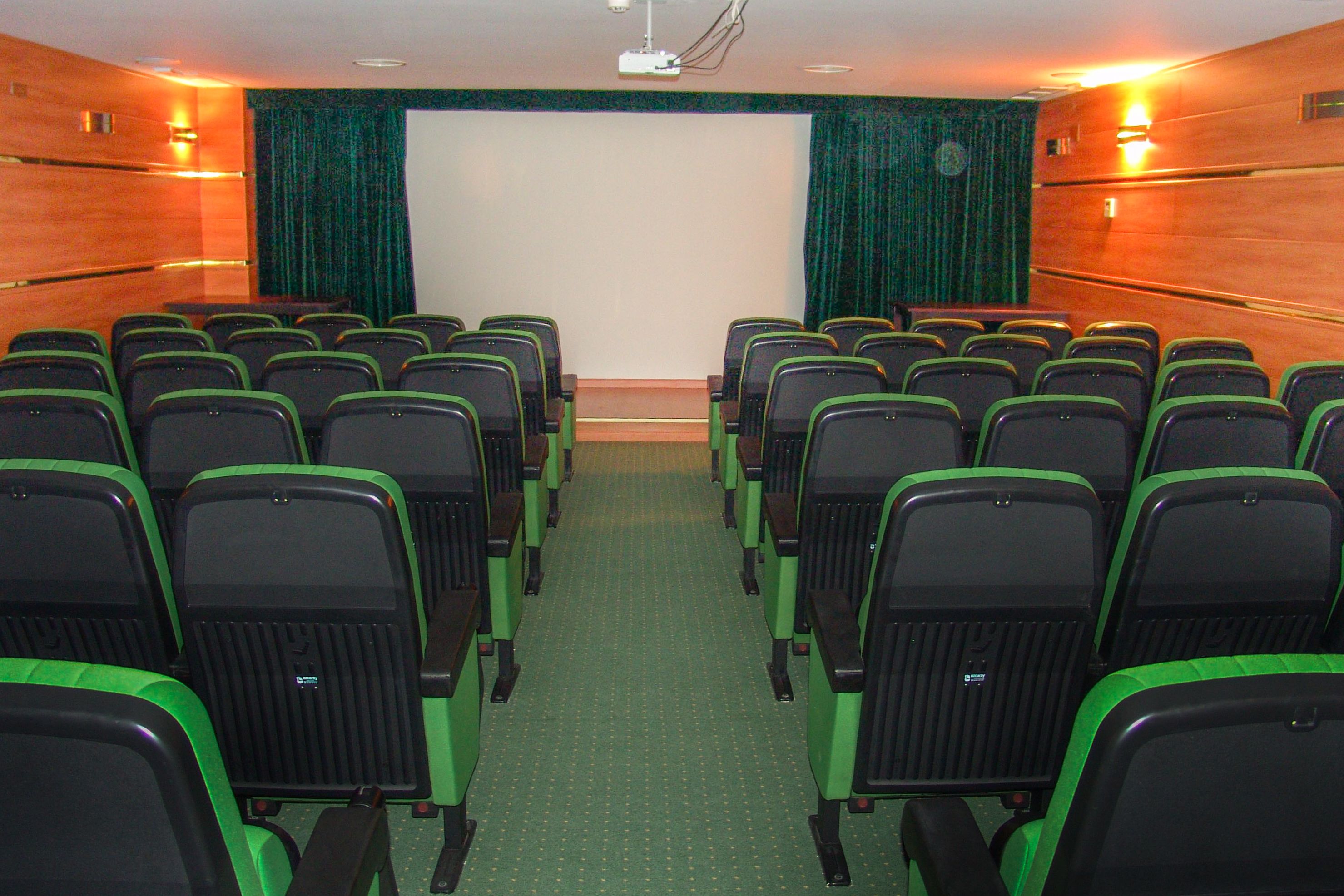 A conference room with rows of green and black chairs, a projector screen, and curtains.