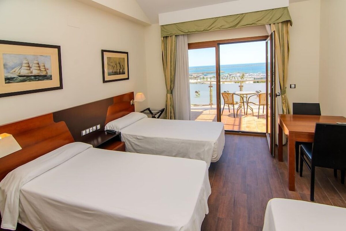 A hotel room with two beds, a dining table, chairs, a desk, and a balcony with a view of the sea.