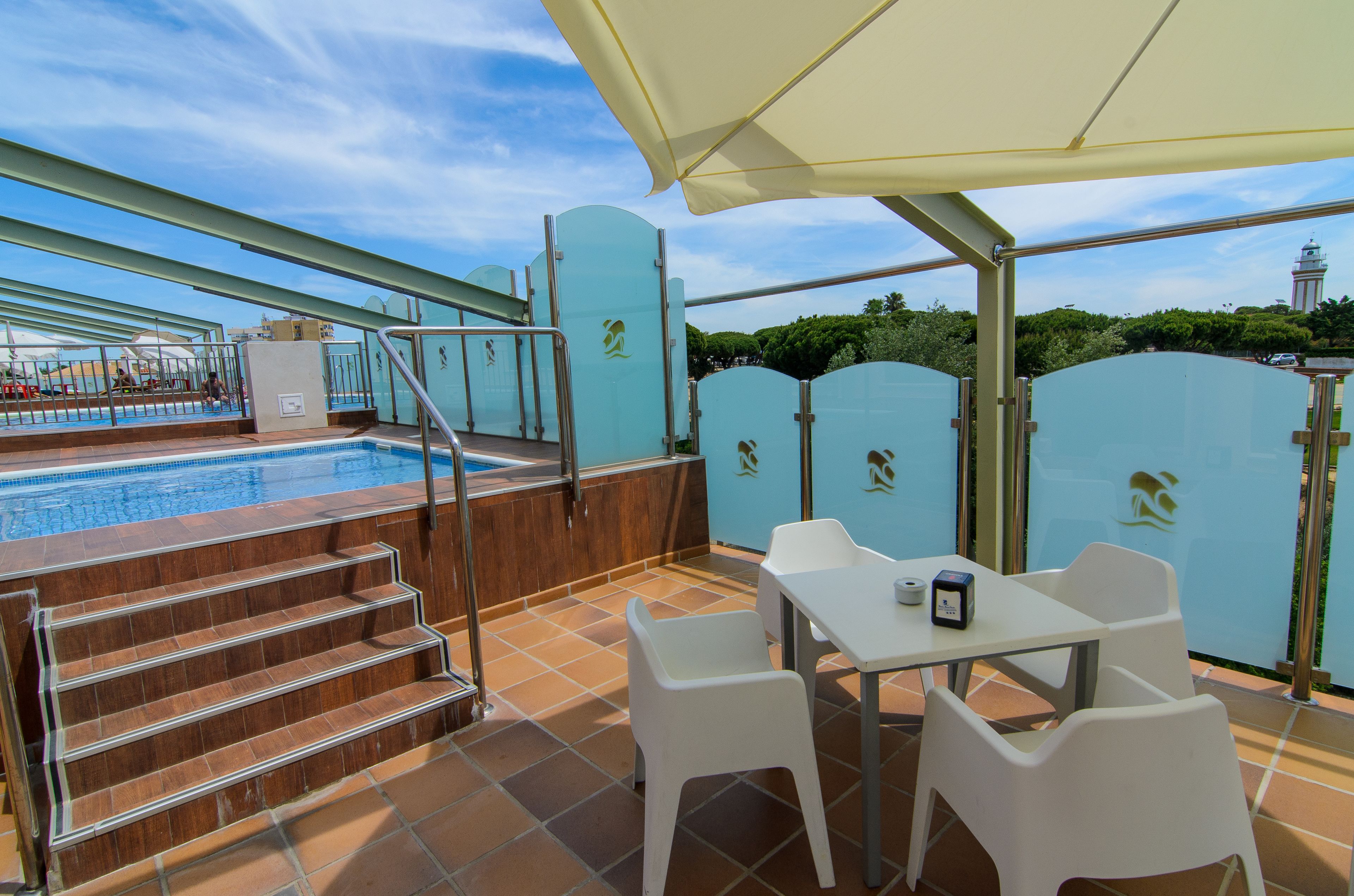 A rooftop pool area with a view of a tower, steps leading to the pool, and a table with chairs under a canopy.