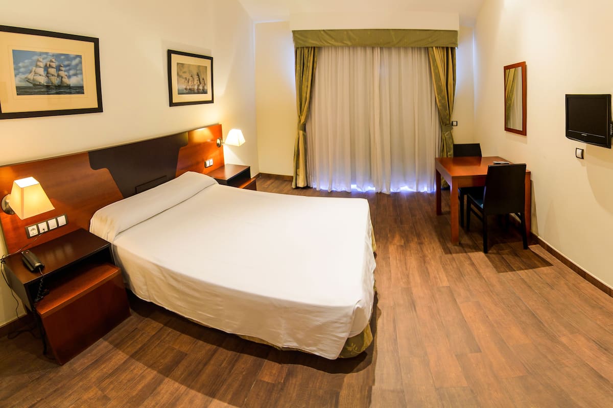 A hotel room with a large bed, bedside tables with lamps, a desk with a chair, a television, and curtains.