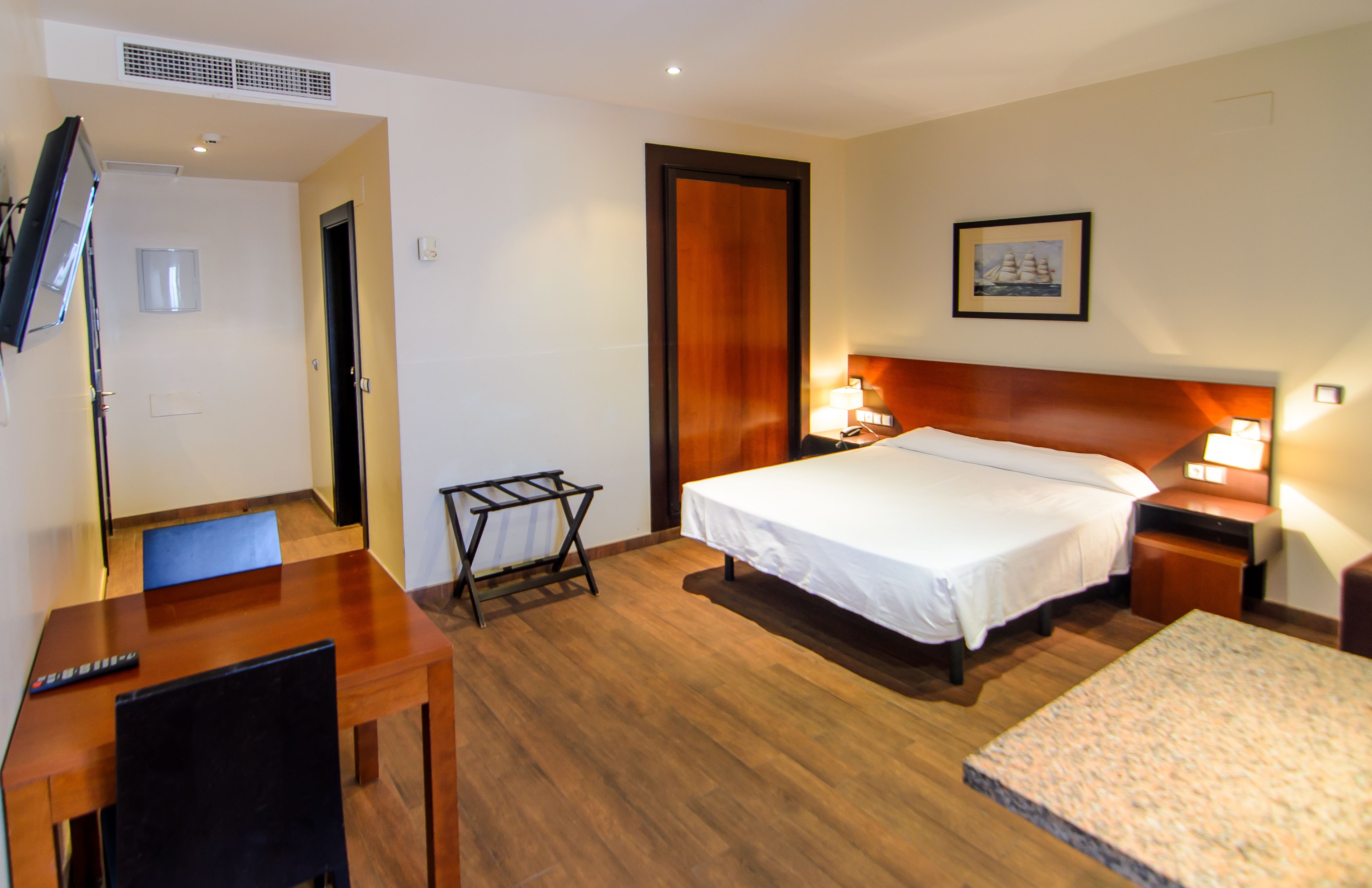 A hotel room with a bed, bedside table, lamp, TV, and a desk with a chair.