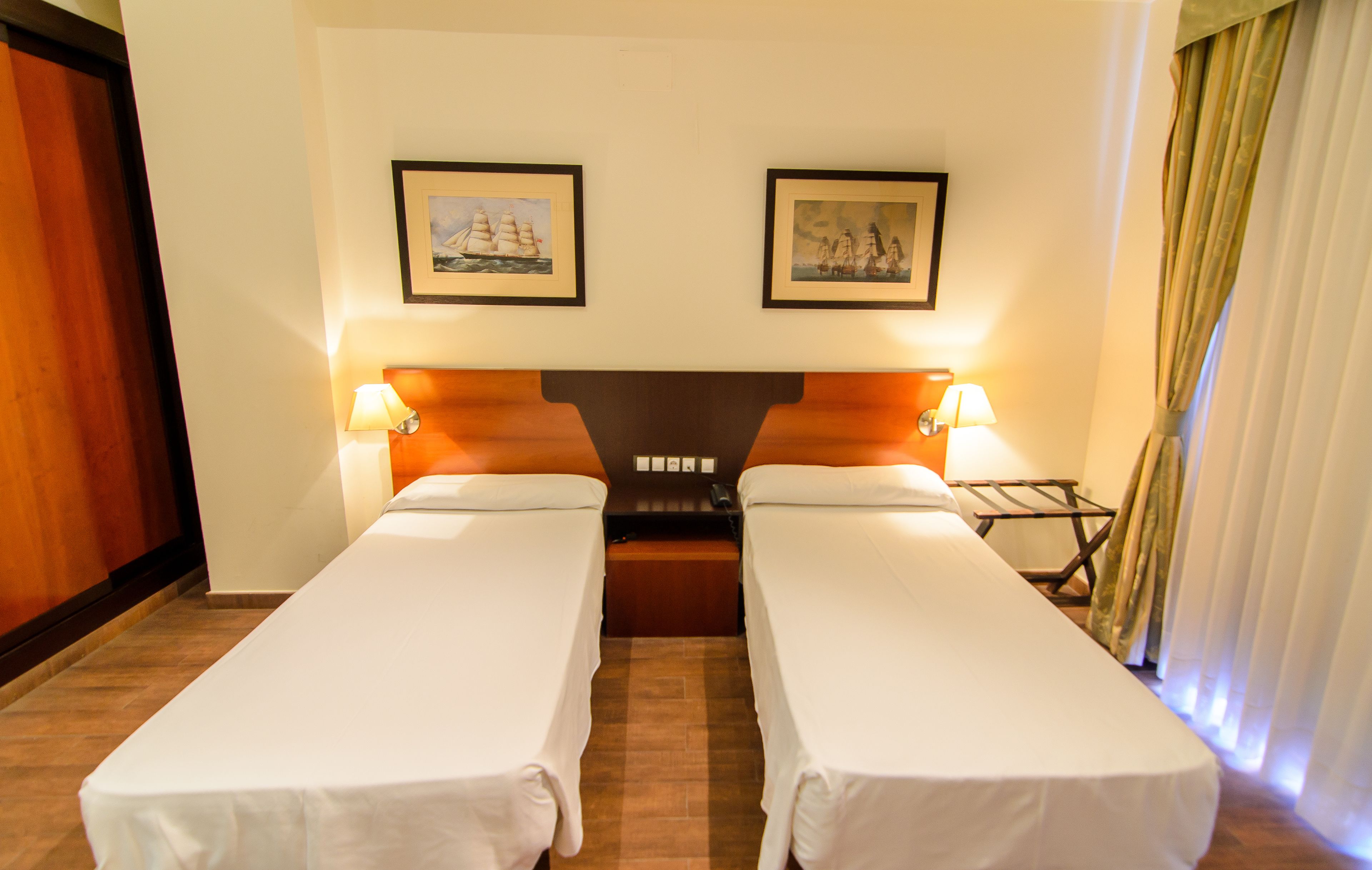 A hotel room with two single beds, a wooden headboard, two framed pictures on the wall, a nightstand with a lamp, and a window with curtains.