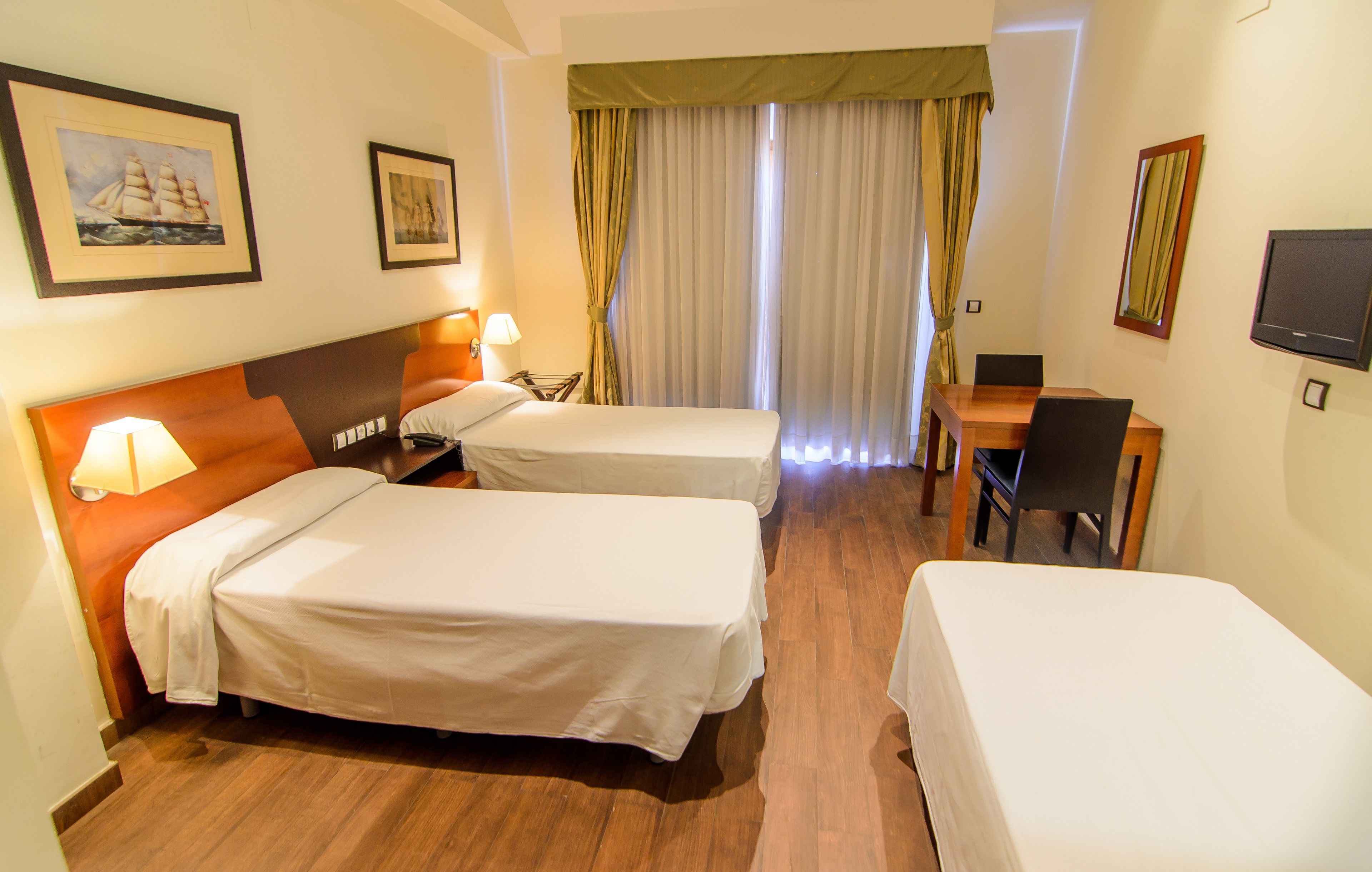 Habitació d'hotel amb dos llits, una taula de treball, una cadira, un televisor i una finestra amb cortines.