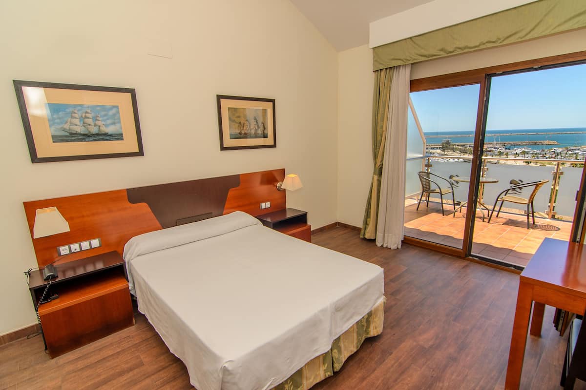 A hotel room with a bed, a desk, a chair, a table, a lamp, and a view of the sea through a balcony.