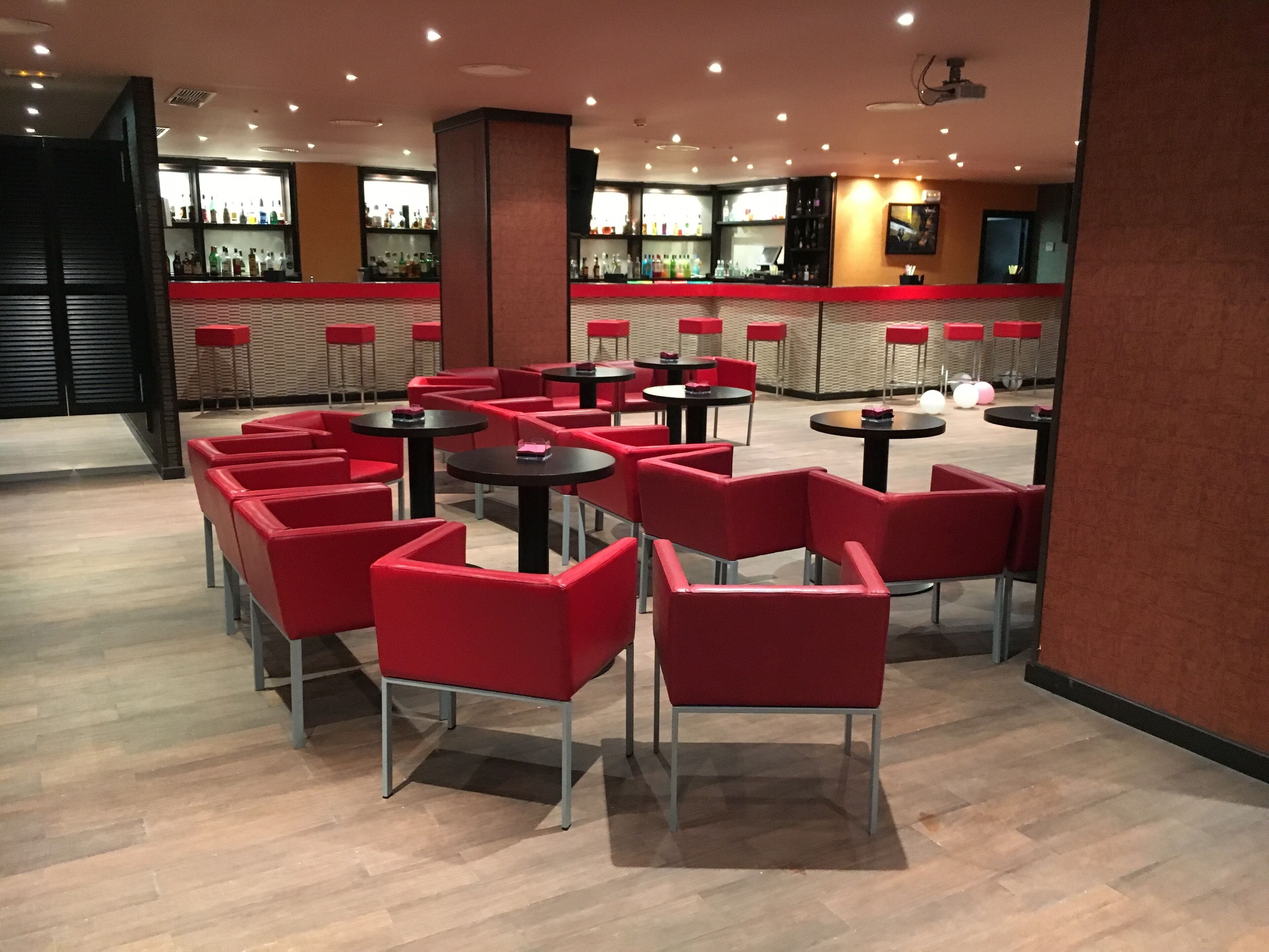Un área de bar moderna con sillas y mesas rojas, una barra con diferentes botellas e iluminación empotrada en el techo.