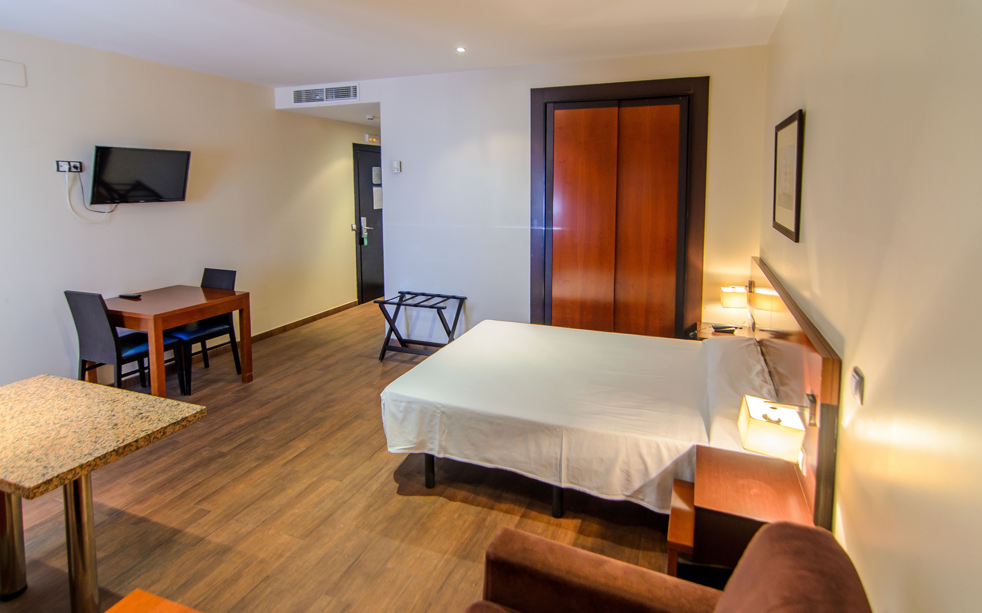 Una habitación de hotel con una cama, un escritorio con silla, un televisor montado en la pared y una puerta.